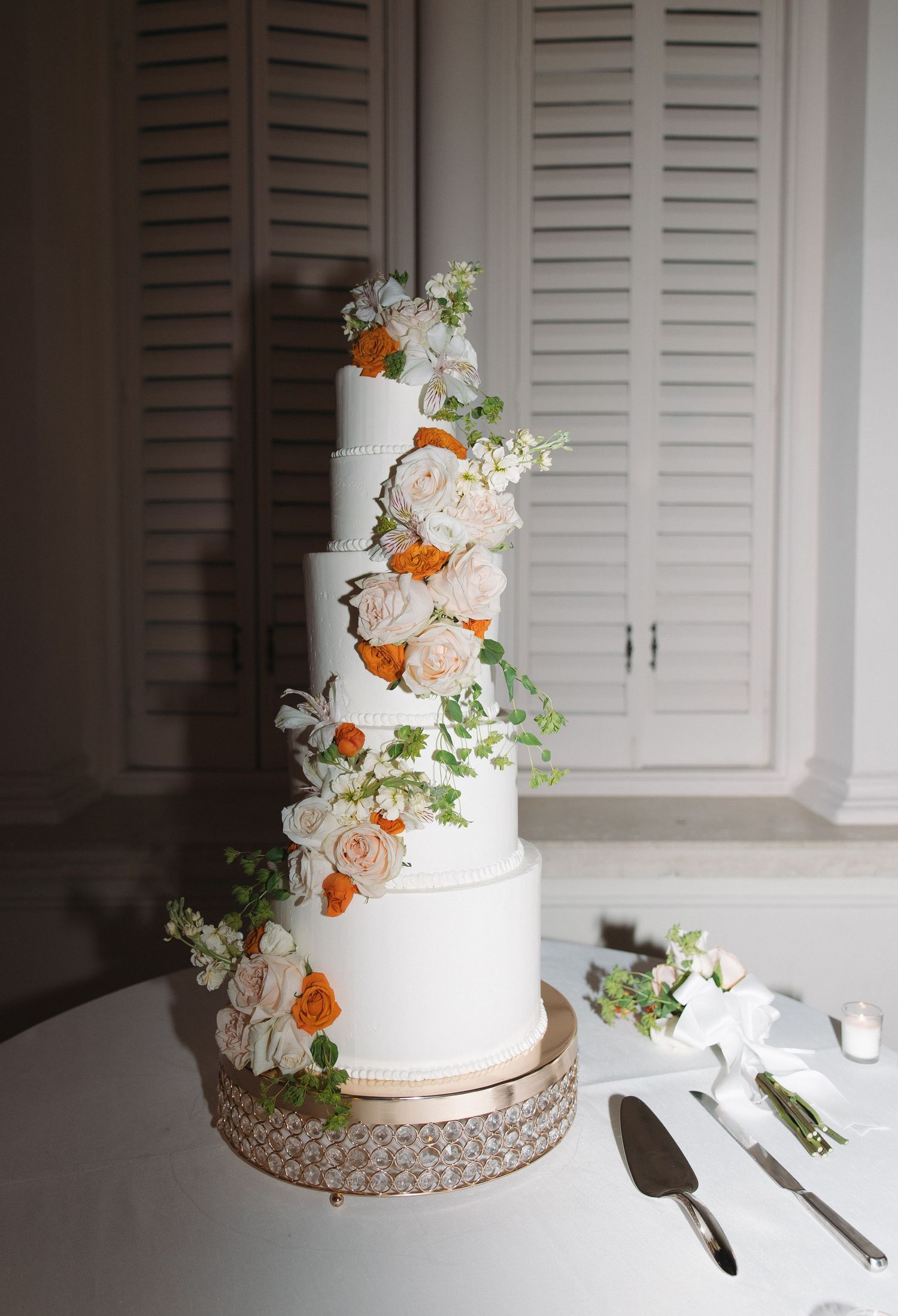 Elegant five-tier white wedding cake decorated with peach and white flowers on a crystal cake stand with serving utensils.