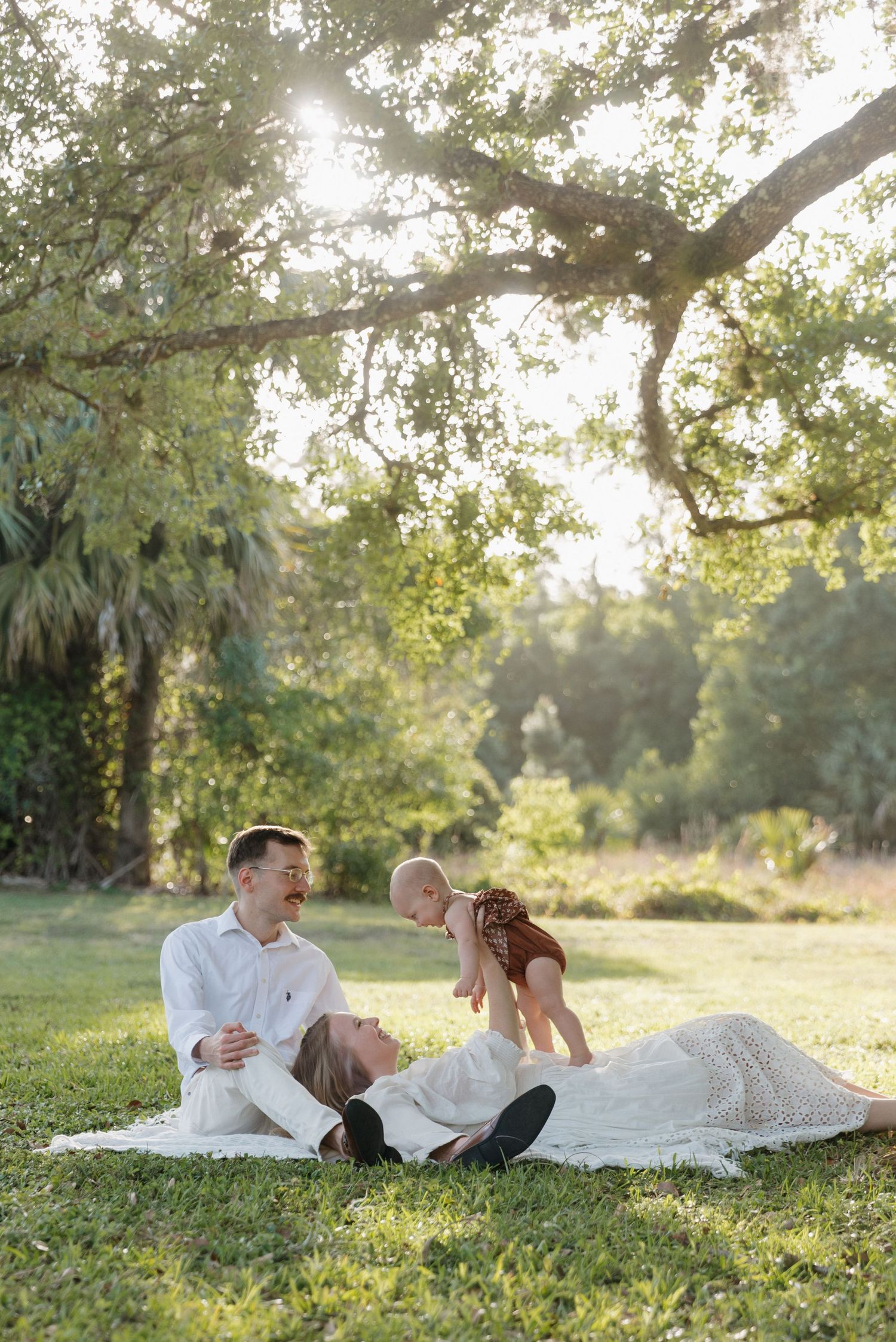 A family relaxes together on a blanket in a sunlit garden with palm trees and oak branches overhead.