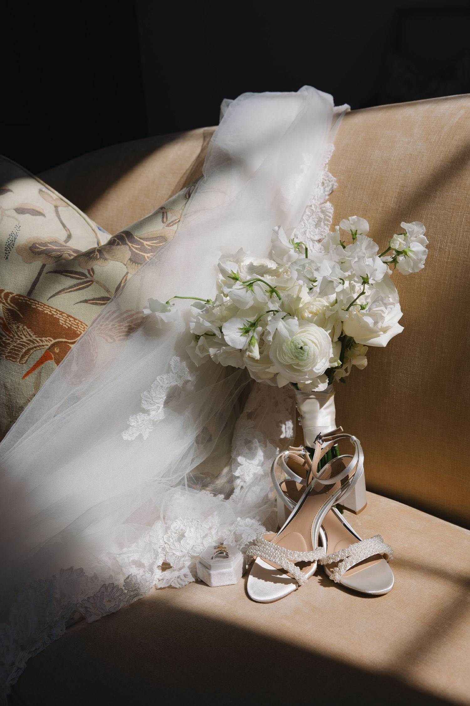 A delicate white bridal bouquet and elegant heels rest on a beige couch in natural light.