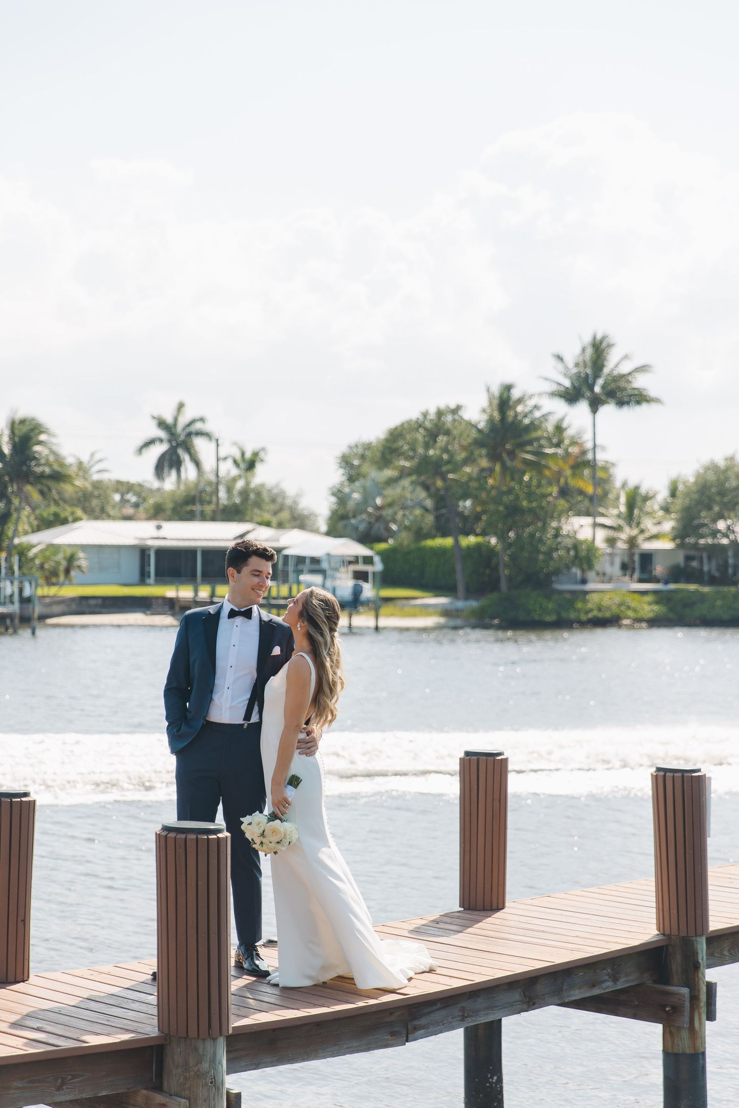 Newlyweds share a romantic moment on a wooden dock overlooking the water with palm trees in the background.