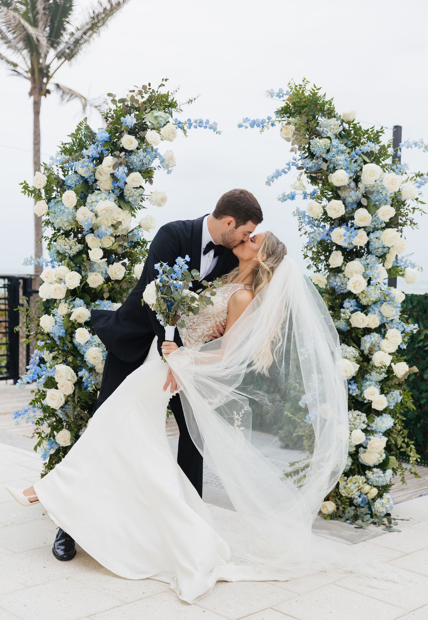 Romantic wedding portraits under floral arch with white roses and blue delphinium flowers.