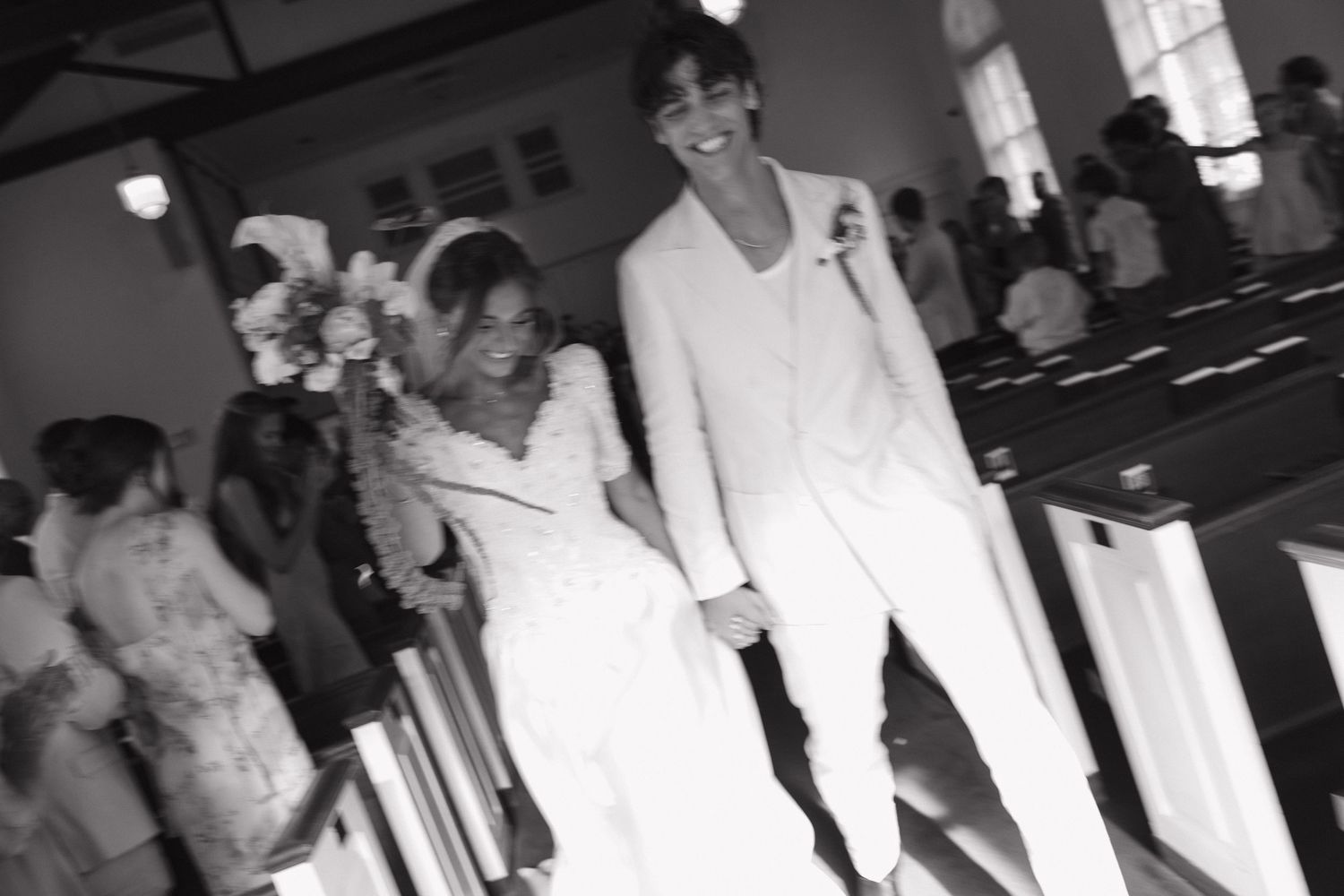 A couple in white formal attire walk down the aisle at their wedding ceremony as guests look on.