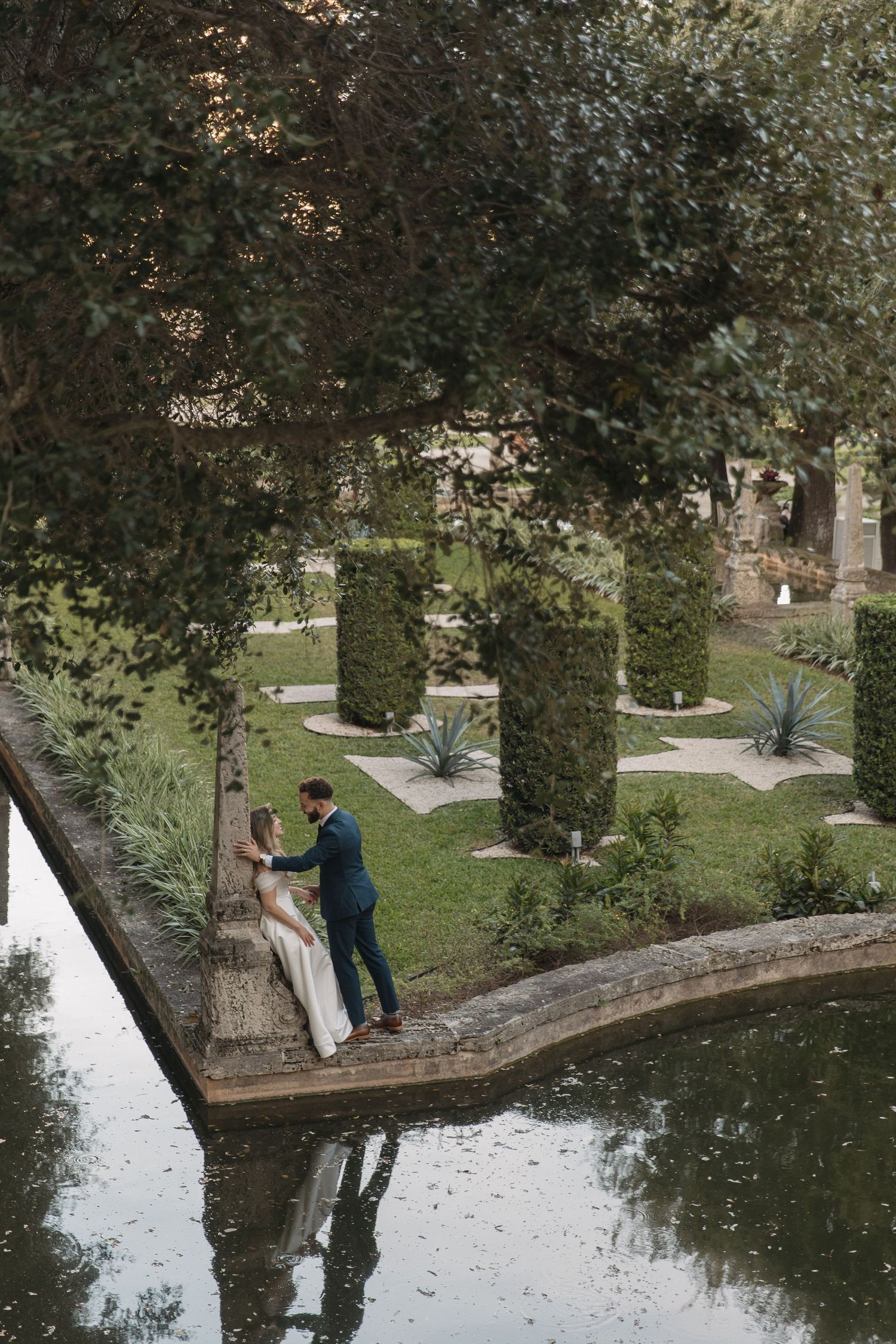 A couple takes photos near a reflective pond in a park setting with trees and greenery in the background.