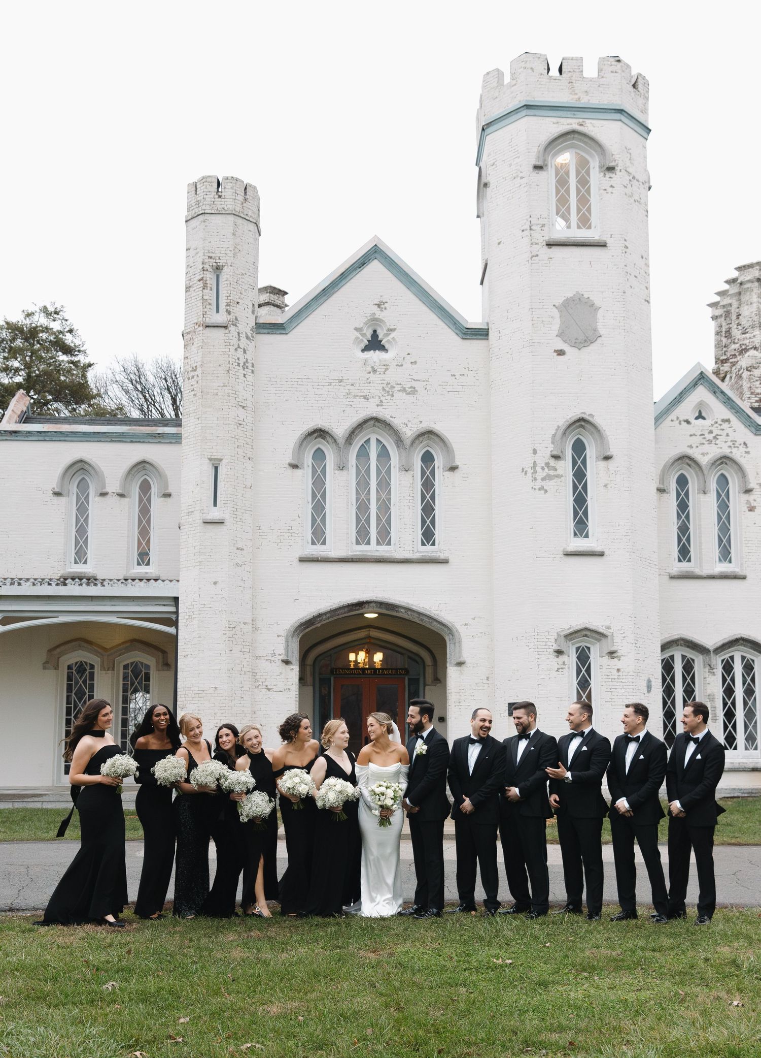 Wedding party in formal attire poses in front of a stunning Gothic Revival white church with multiple archways and towers.
