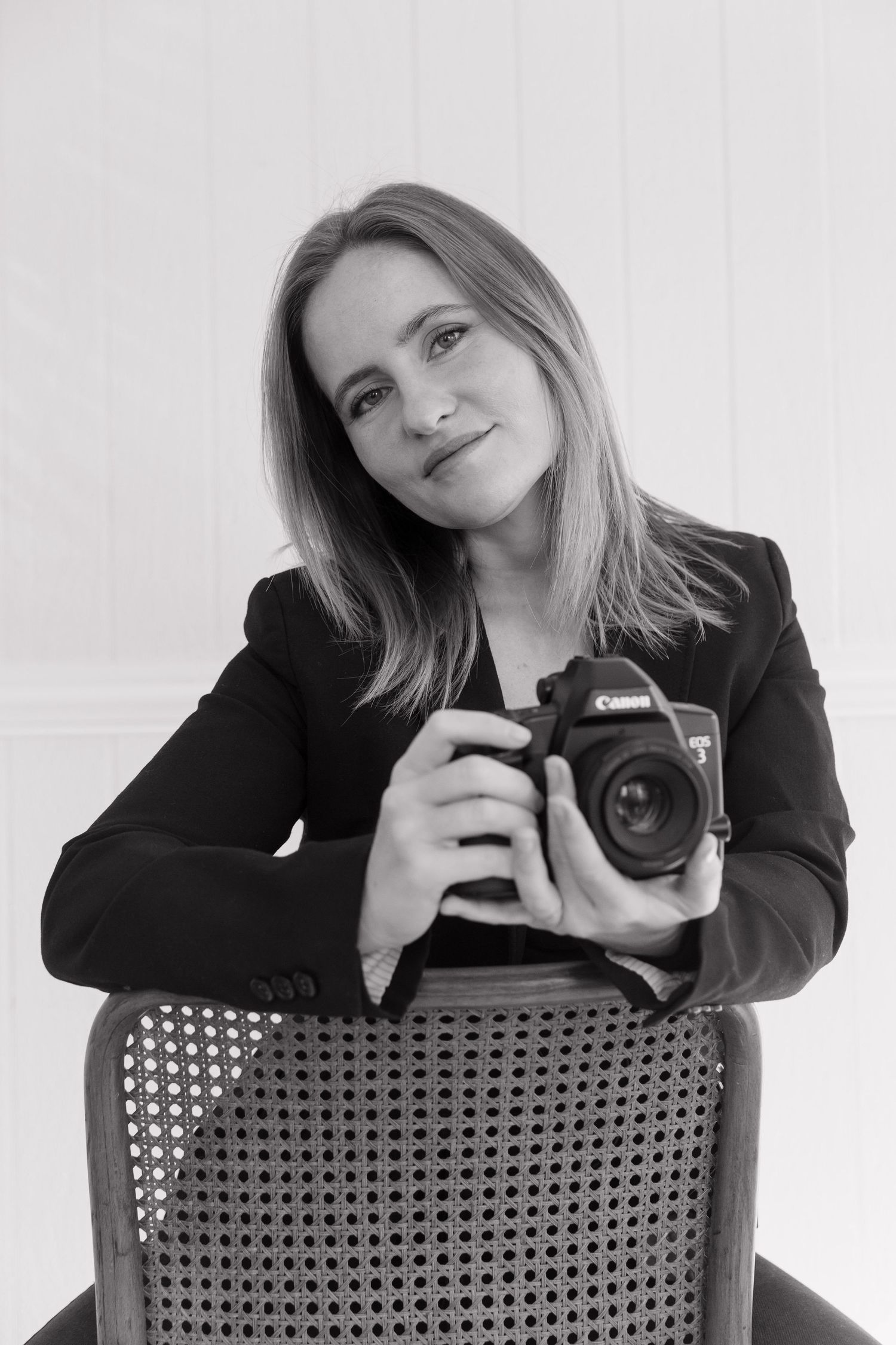 A professional in a black blazer poses with a Canon camera while seated on a cane chair.