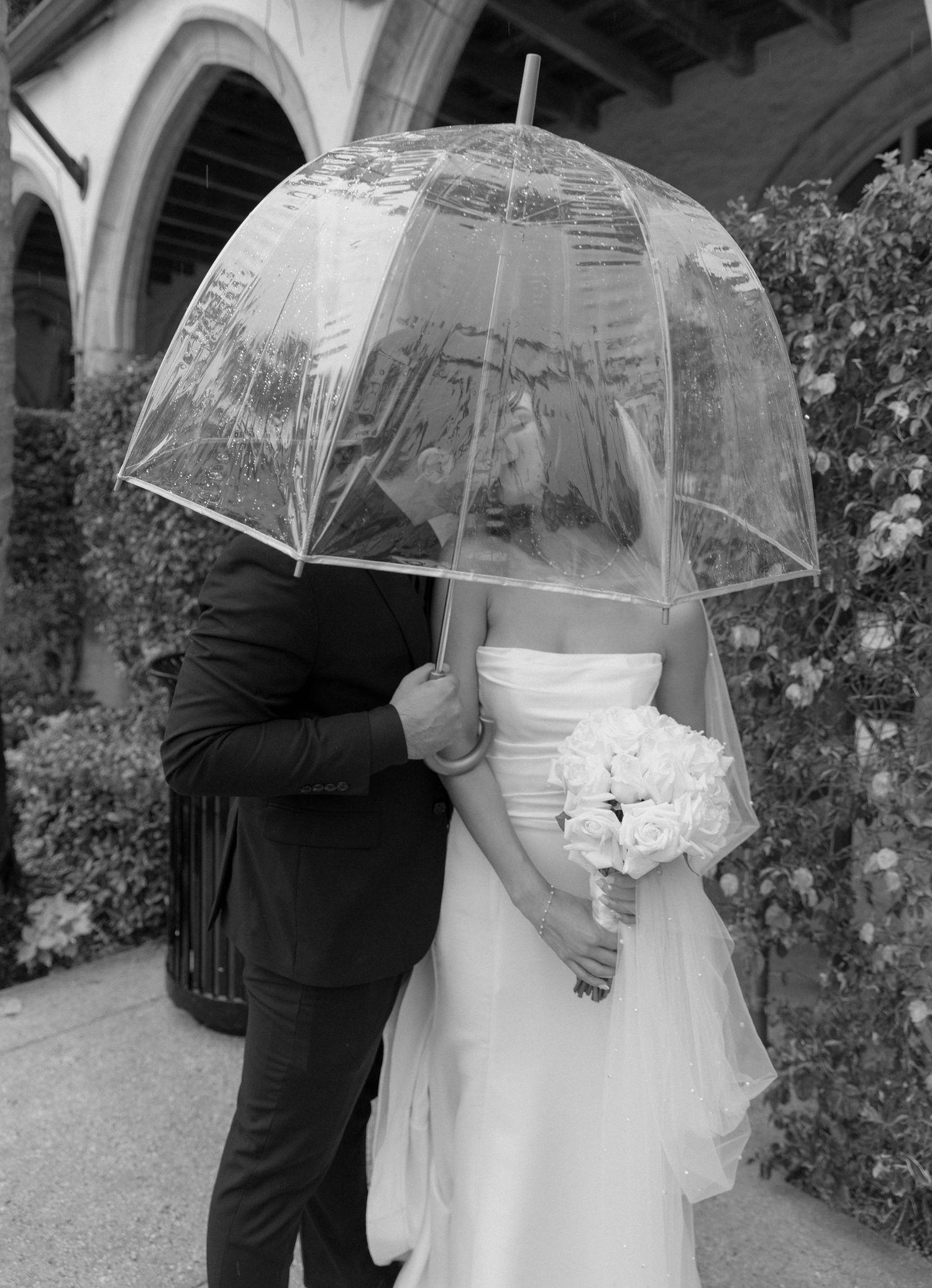 Romantic black and white photo of couple under clear umbrella at wedding venue.