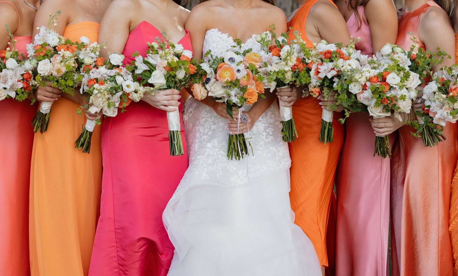 A row of bridesmaids in alternating coral orange and hot pink dresses hold matching white floral bouquets.