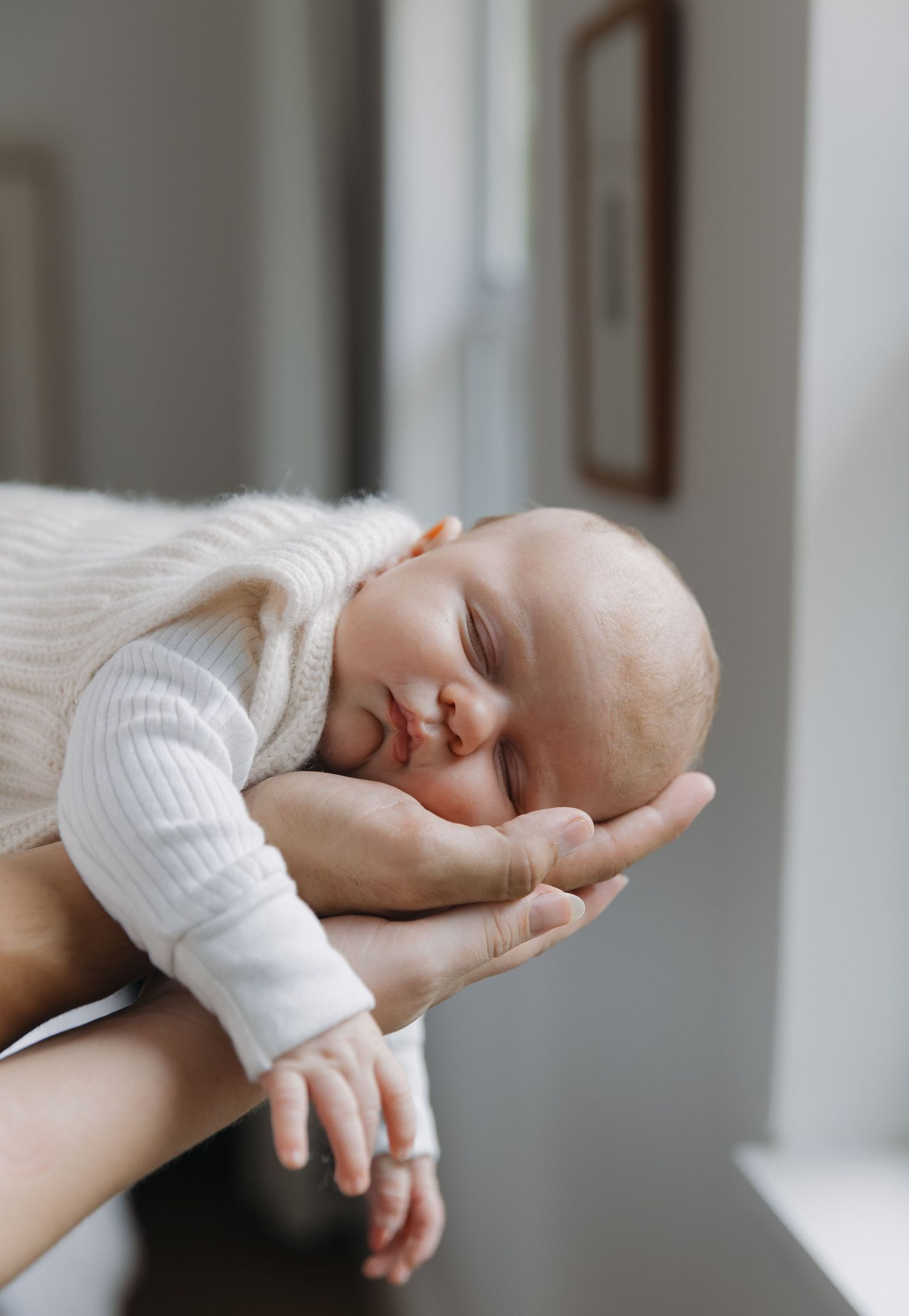 A sleeping newborn baby rests peacefully in gentle hands against a soft white background.