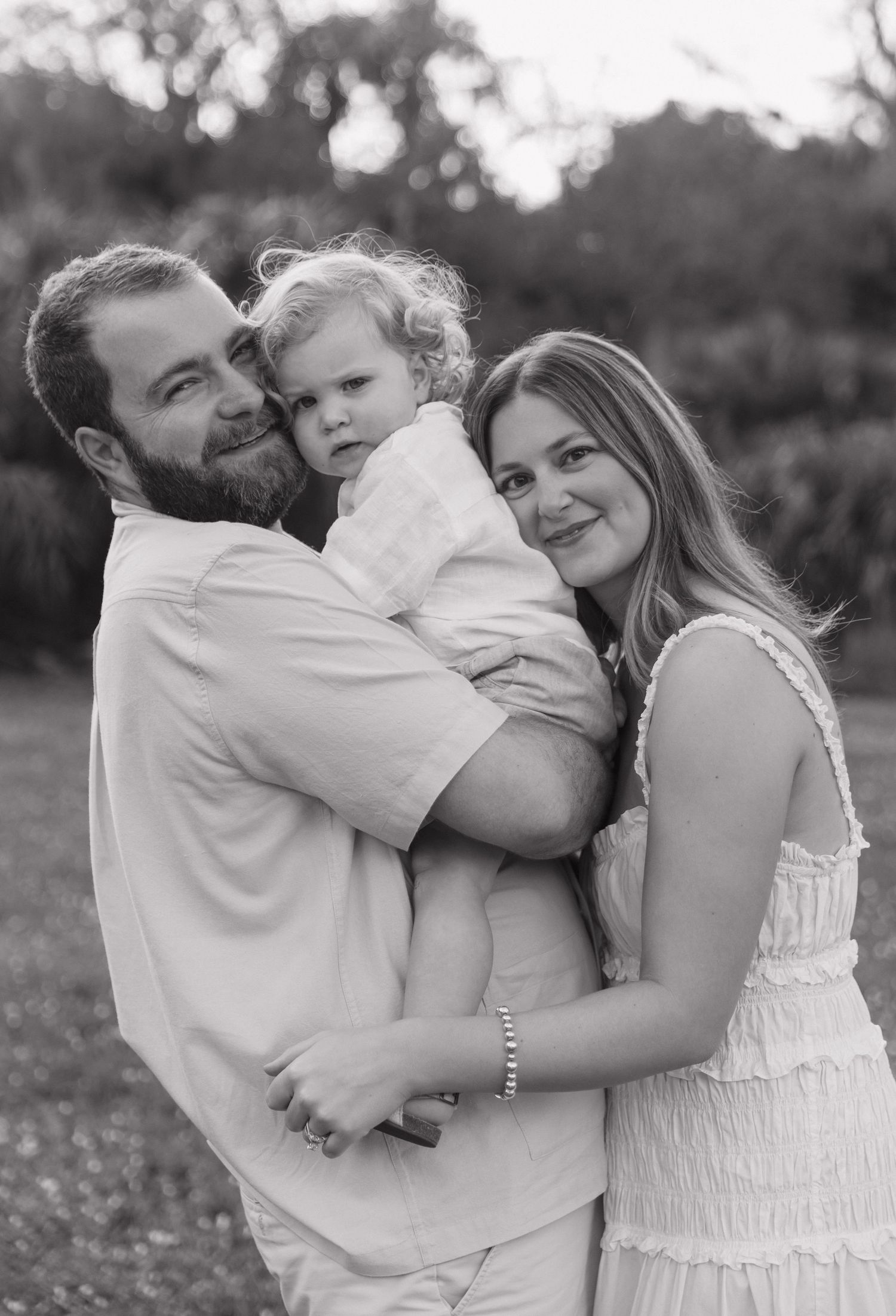 Black and white family portrait of bearded man and woman holding toddler outdoors among trees.