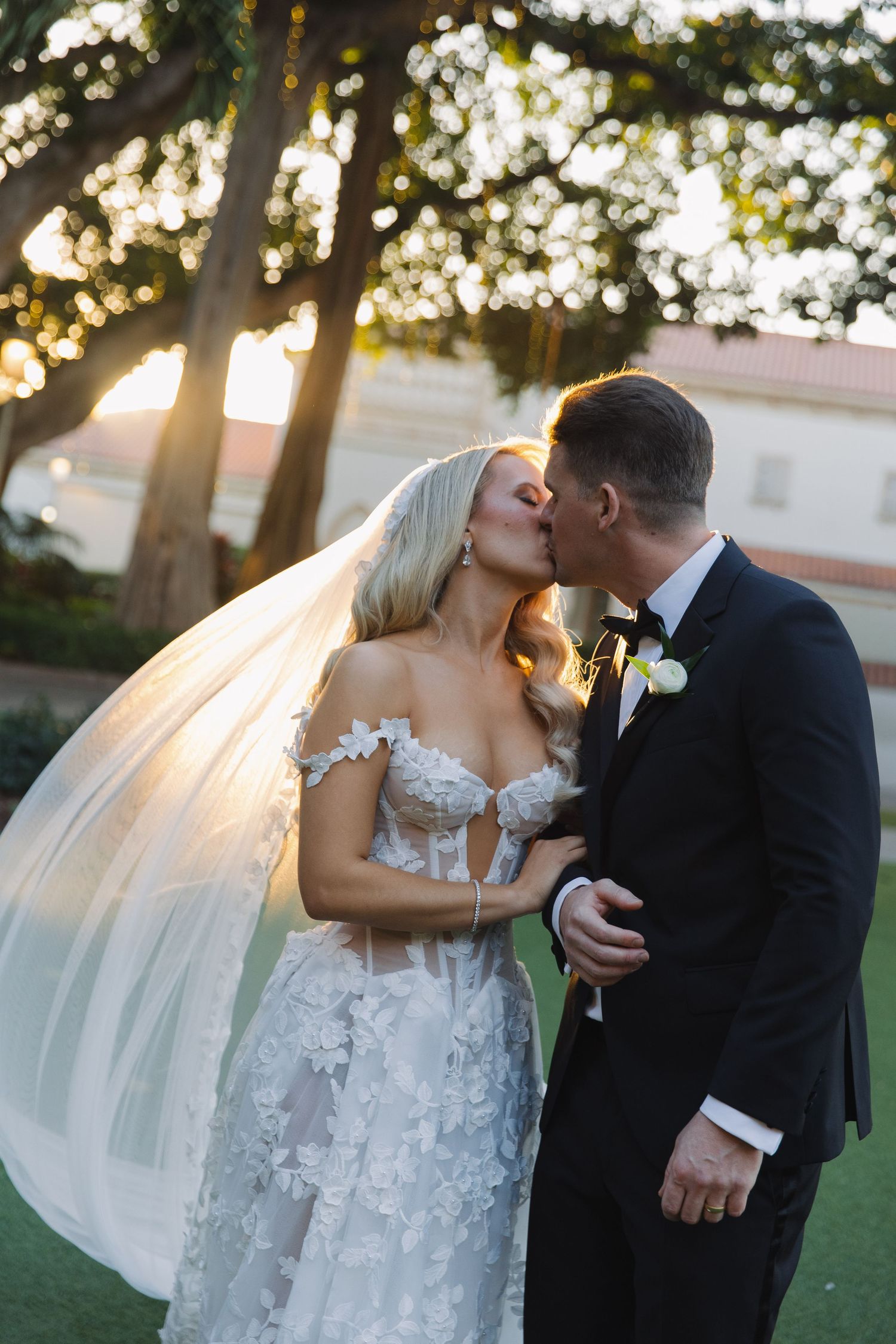 A couple shares a romantic kiss at sunset during their outdoor wedding ceremony, with golden light filtering through trees.