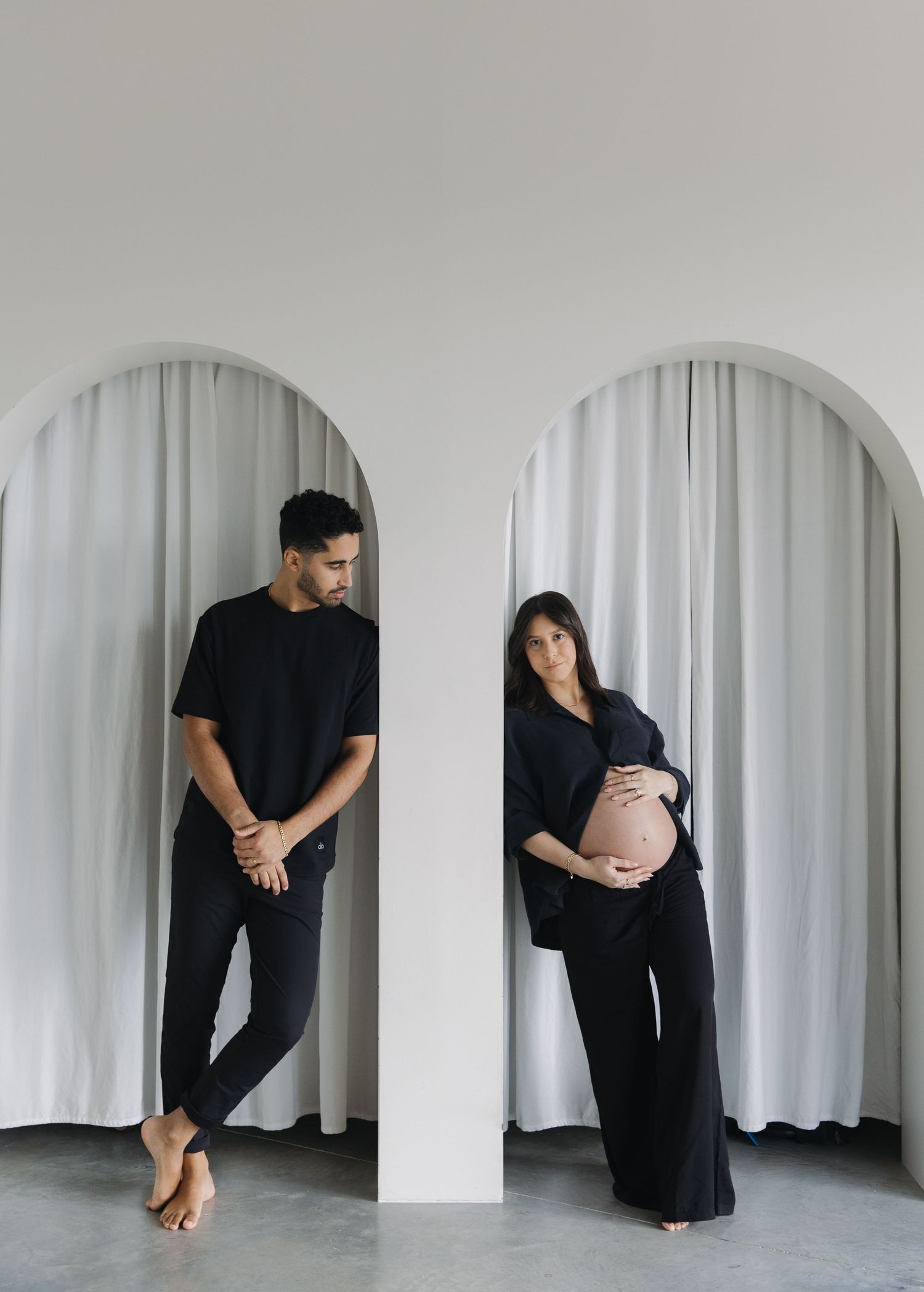 Minimalist maternity photo with couple in black outfits standing in separate white archways against light background.