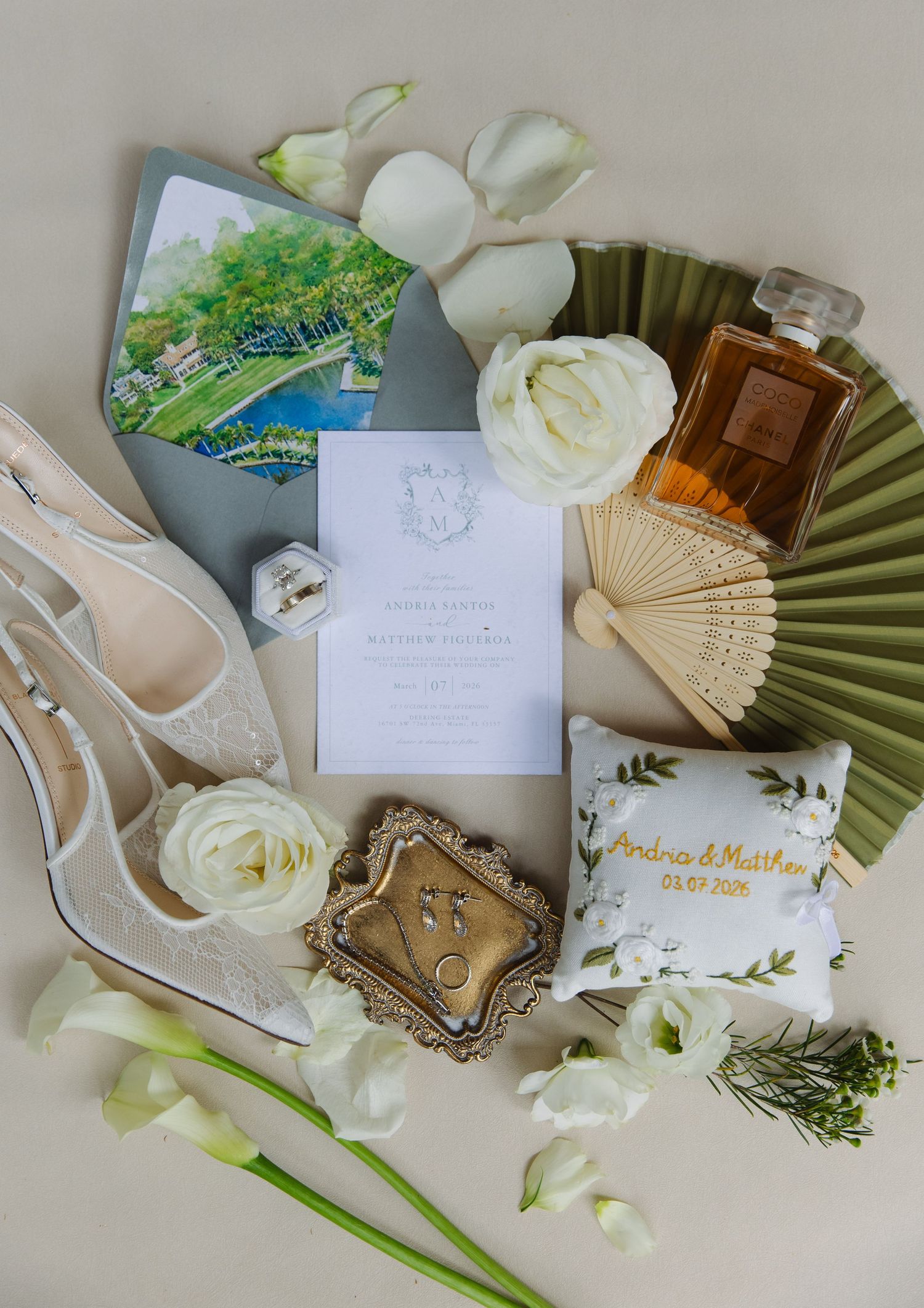 Wedding details flatlay with white roses, invitation cards, heels, perfume bottle and decorative fans arranged elegantly.