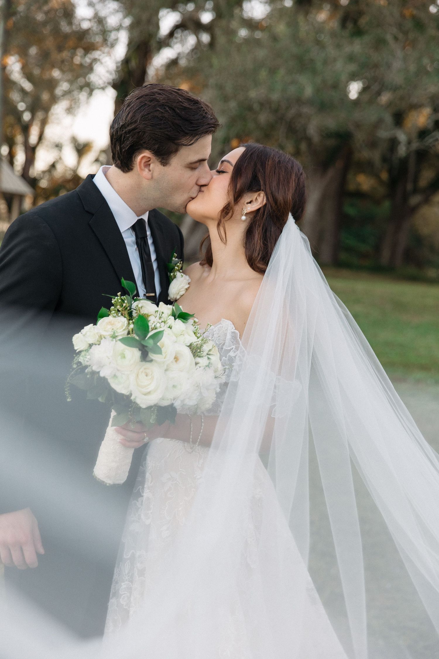 Romantic newlyweds share an intimate kiss outdoors while holding a white rose bouquet.