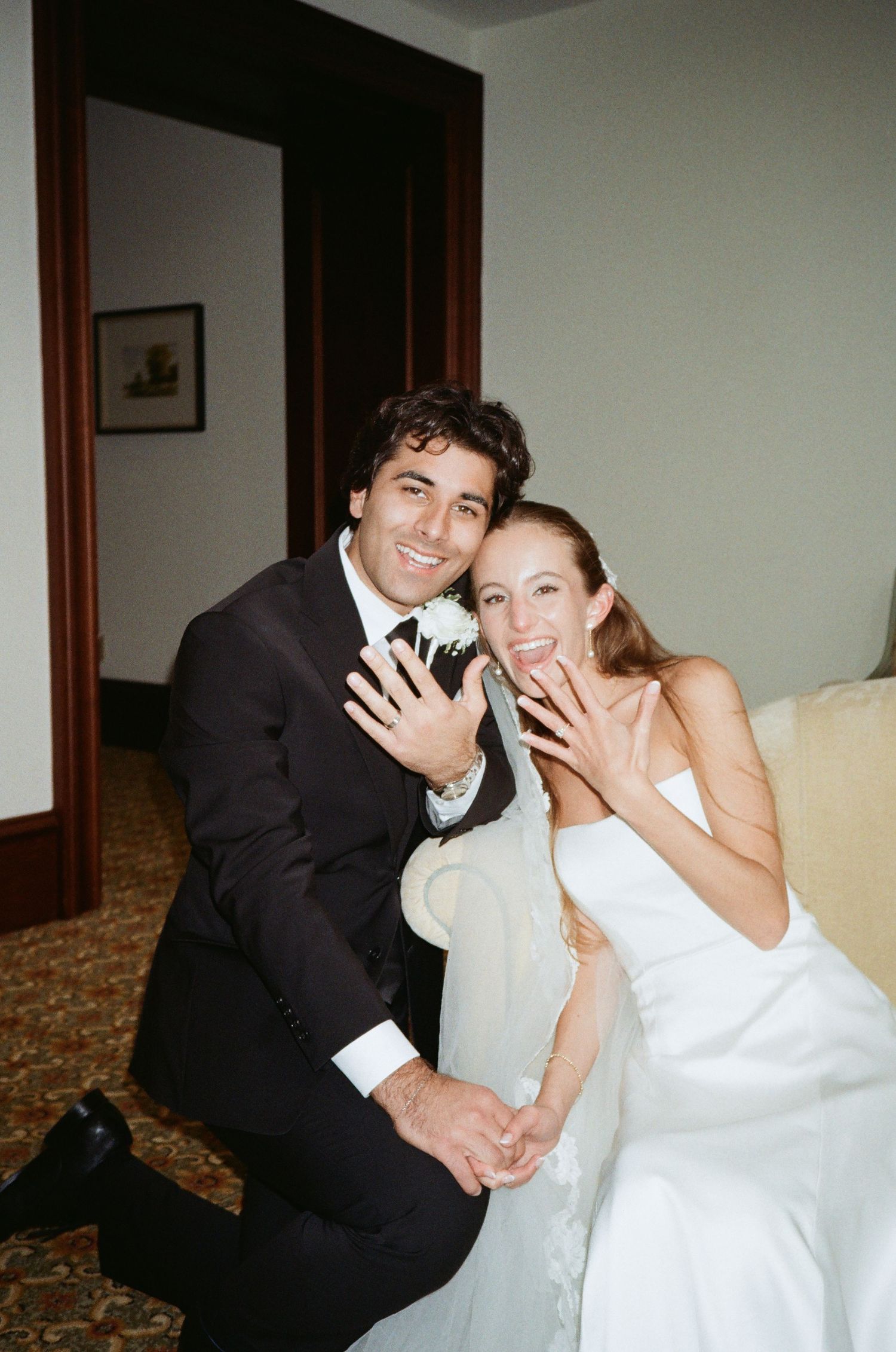 A playful wedding couple poses together indoors with wooden trim and white walls in the background.