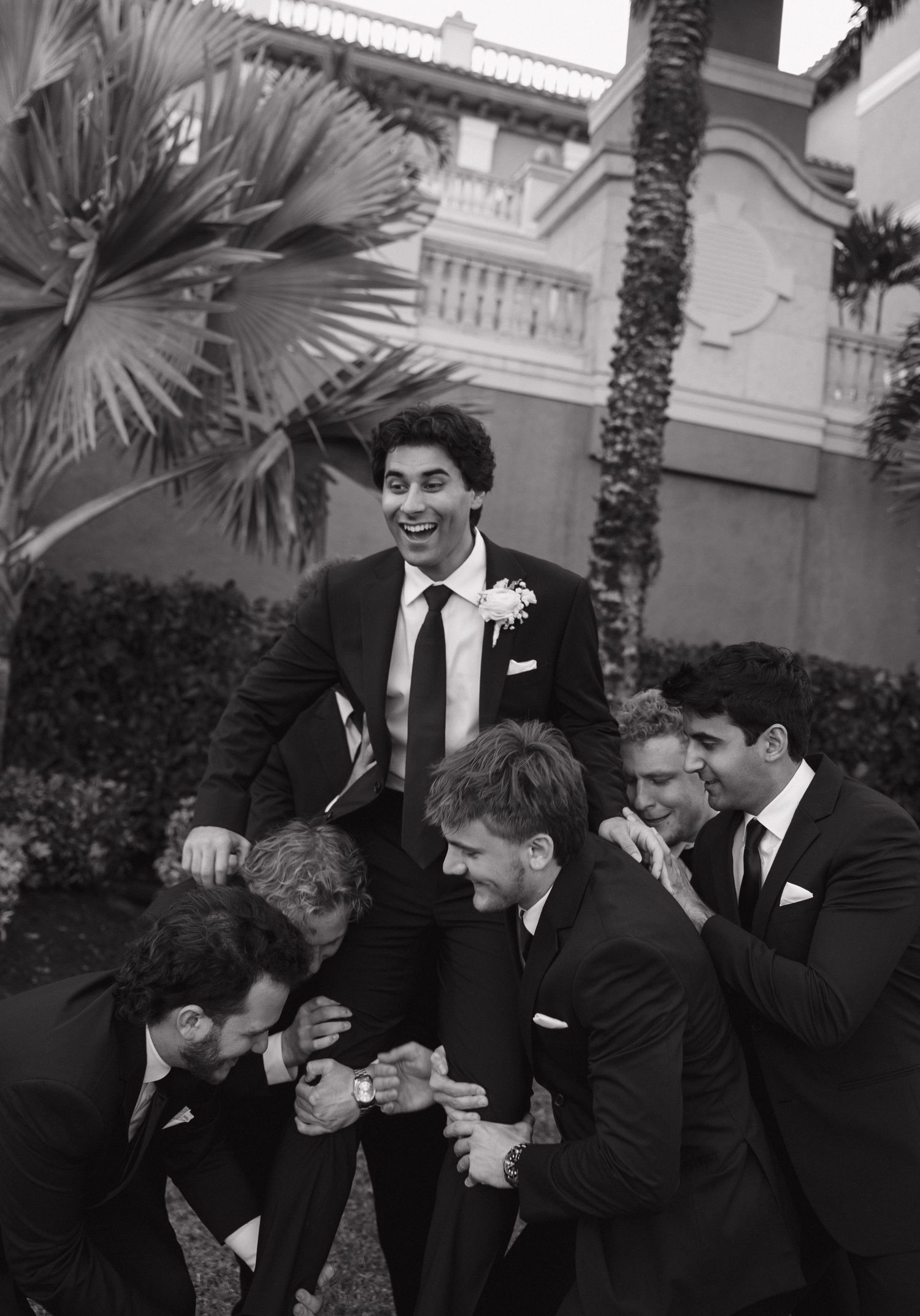 Black and white photo of groomsmen laughing together outdoors by palm trees at a formal event.
