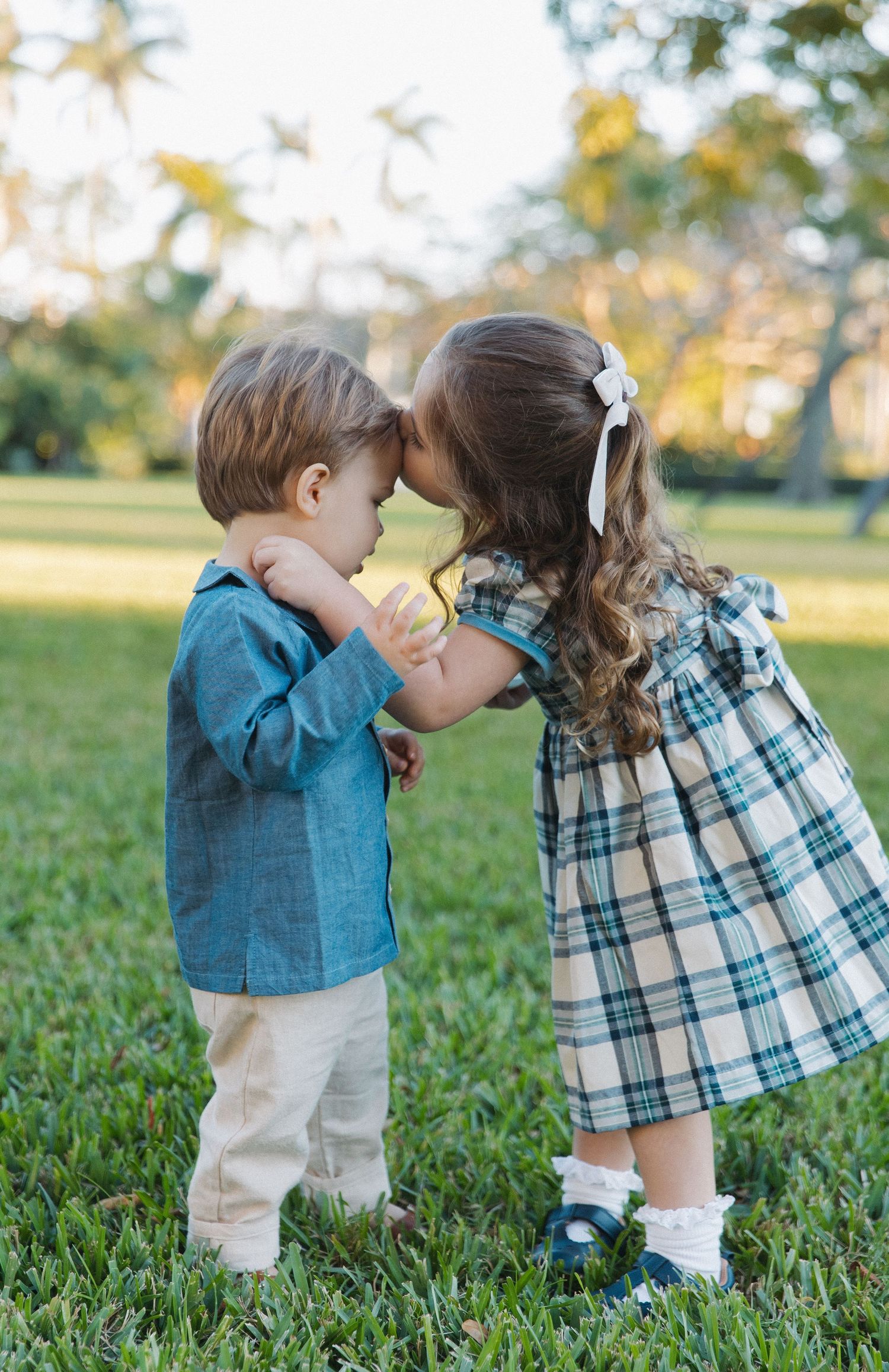 Two young children in coordinating blue and plaid outfits share a sweet moment together in a grassy field at sunset.