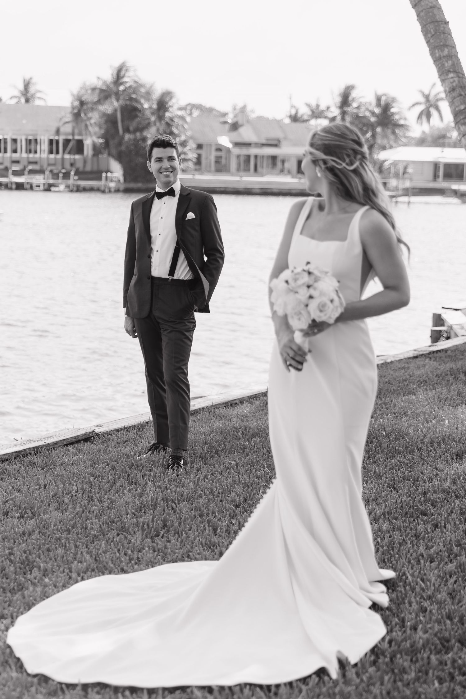 Black and white photo of newlyweds at a waterfront location, with palm trees and tropical houses in the background.