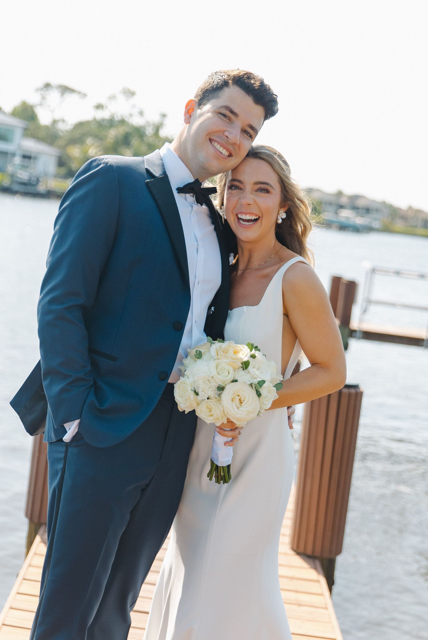 Wedding couple smiling on a waterfront dock with boats in background.