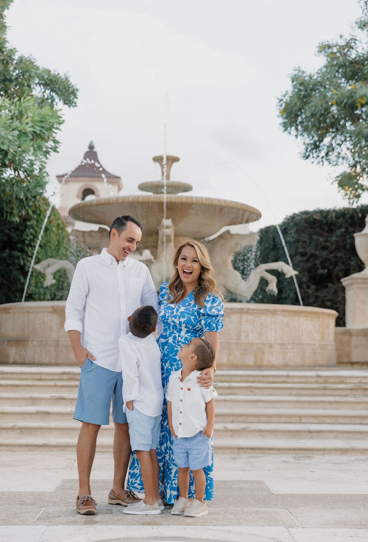 Family wearing coordinated blue and white outfits poses playfully near a stone fountain in an outdoor setting.