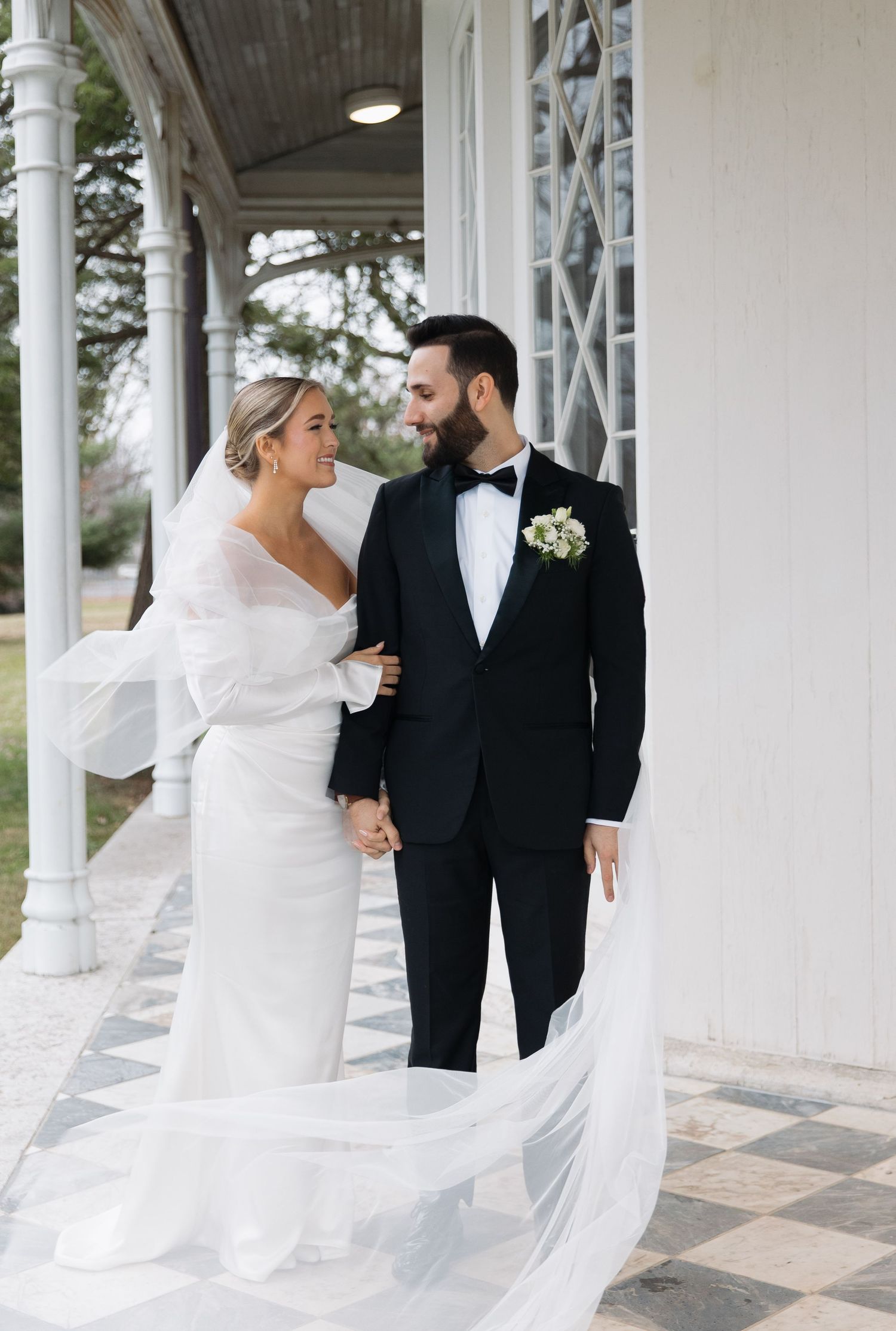 A romantic wedding couple stands on a white columned porch as the bride's veil flows elegantly in the breeze.