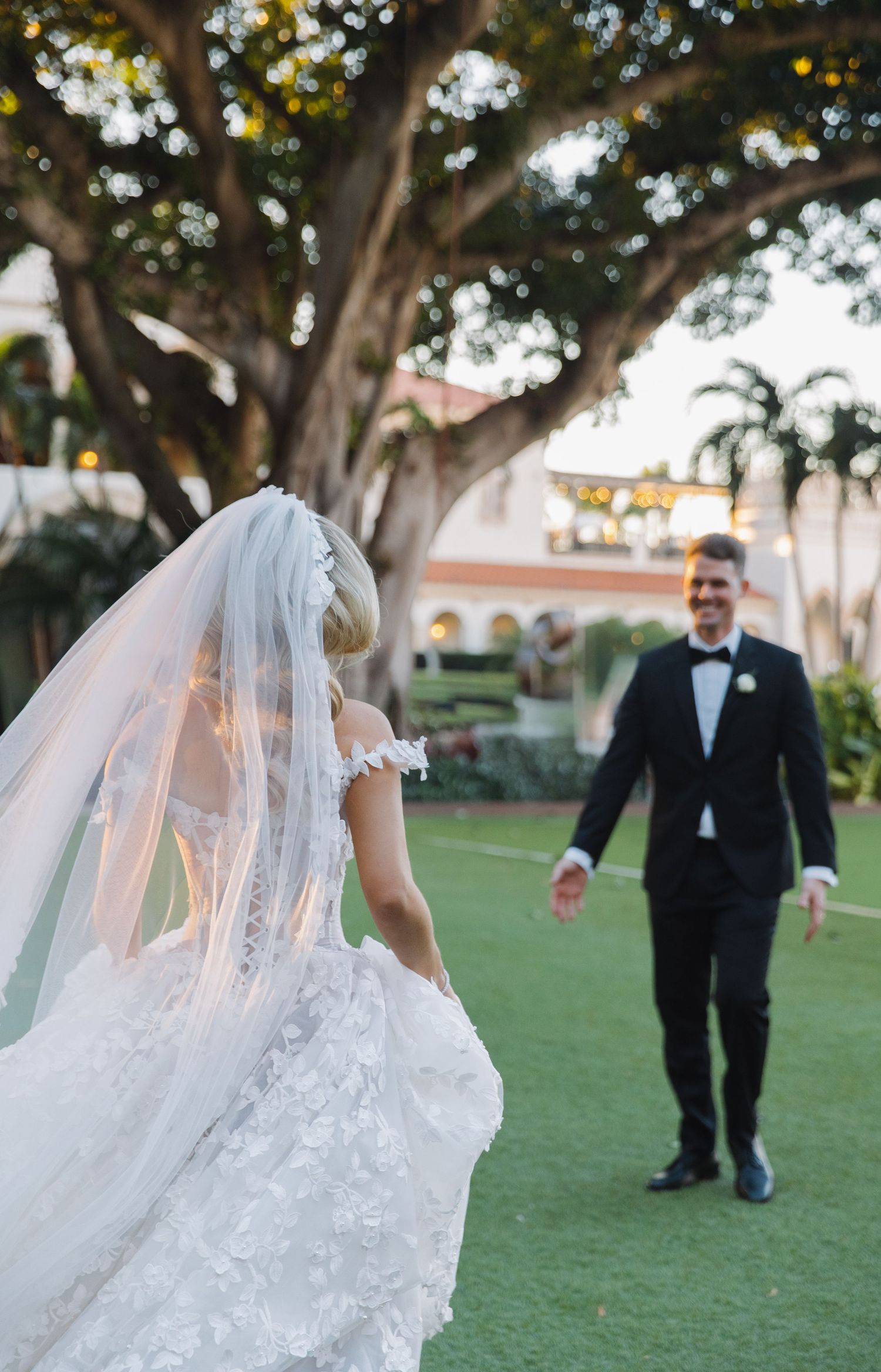 A bride in a flowing lace wedding dress with veil walks across a grassy lawn while a suited figure follows behind.