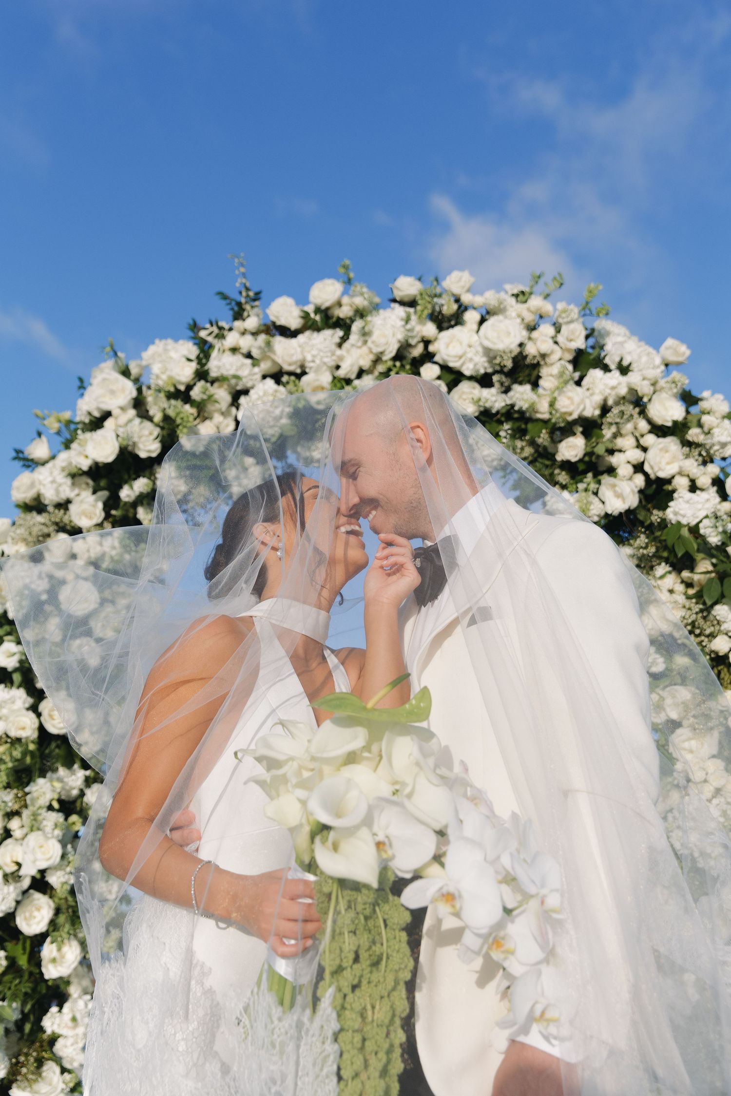 Romantic wedding ceremony sequence under white floral arch with flowing veil and white roses.