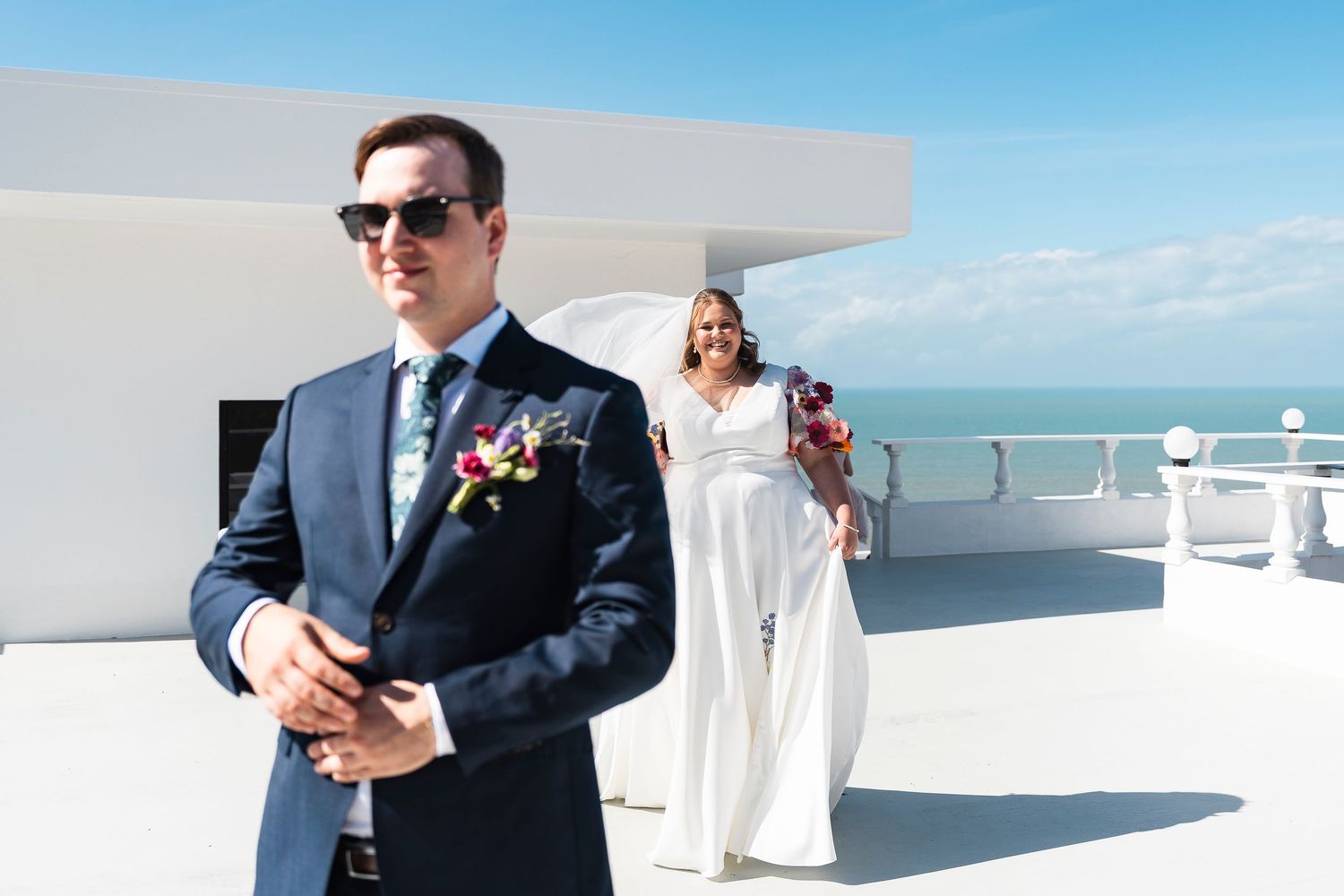 Groom in navy suit stands on rooftop terrace as bride in white gown approaches with bouquet, ocean view behind them.