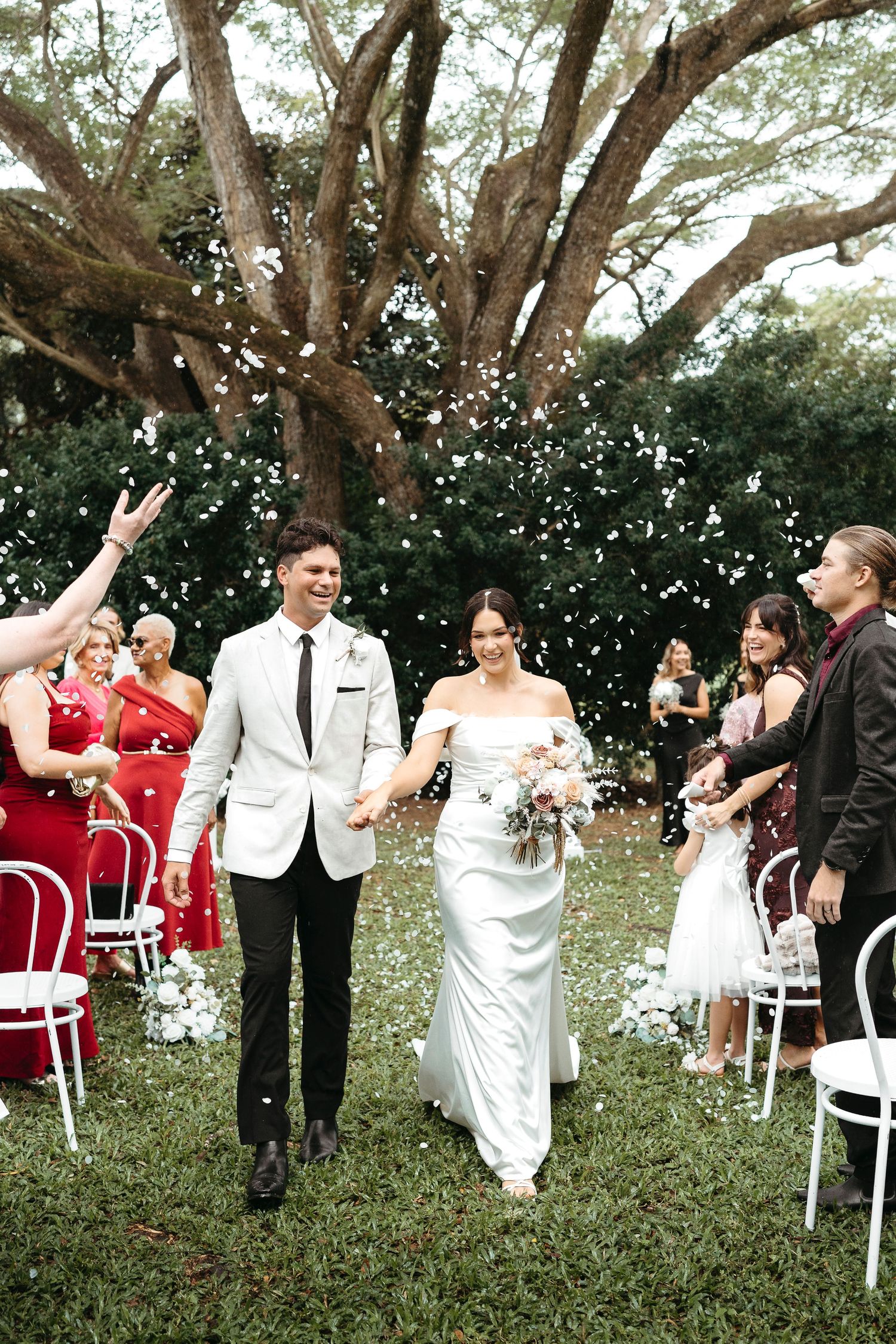 Newlywed couple walks down outdoor aisle as guests toss white confetti petals, bride in white gown holding bouquet.