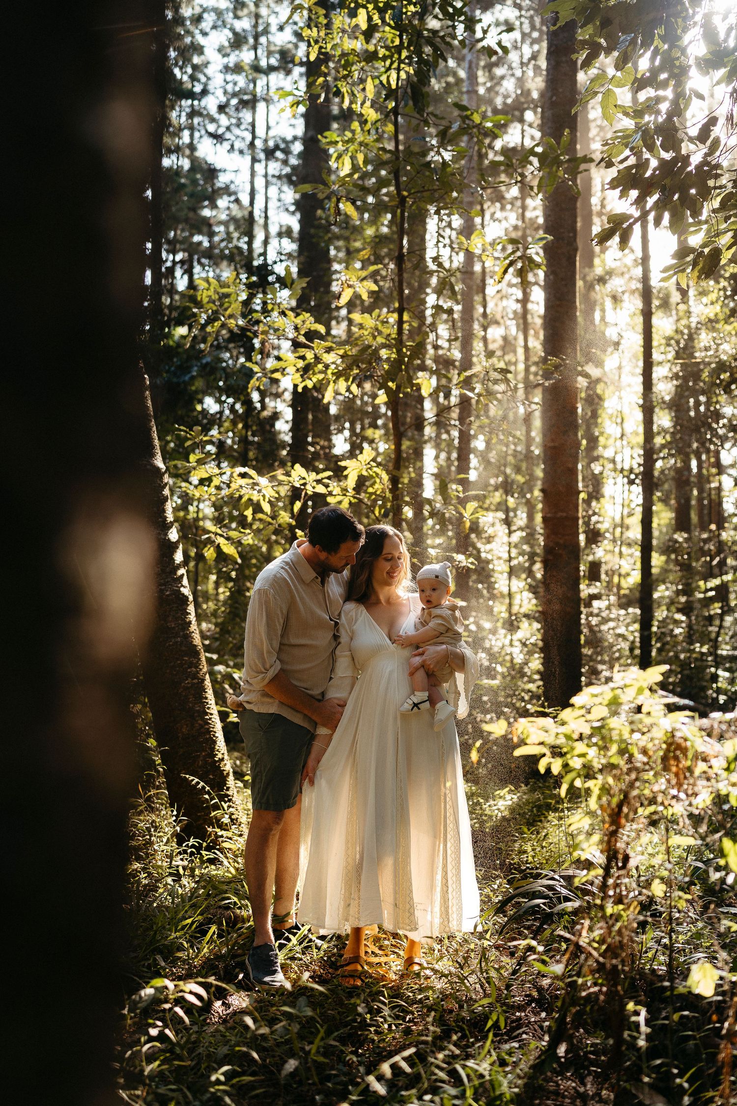 A family with a newborn baby shares a tender moment in a sunlit forest, mother in a flowing white dress.