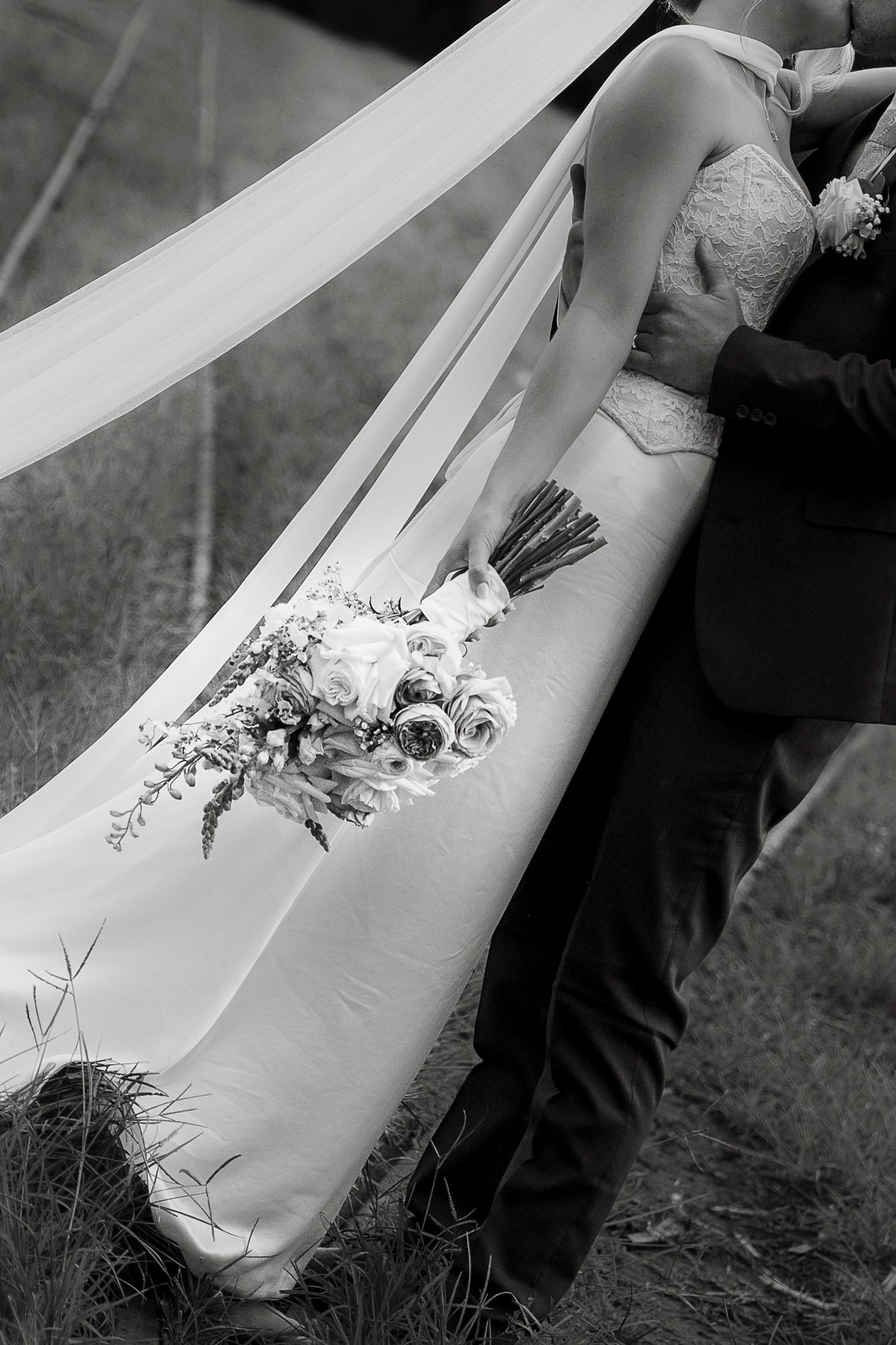 Black and white wedding photo of bride and groom embracing outdoors, with flowing veil and bridal bouquet of roses.