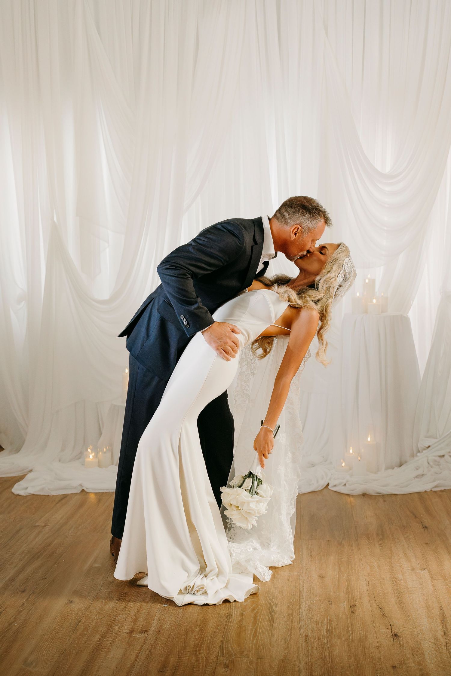 Groom dips bride in sleek white gown for a kiss before elegant draped white backdrop.