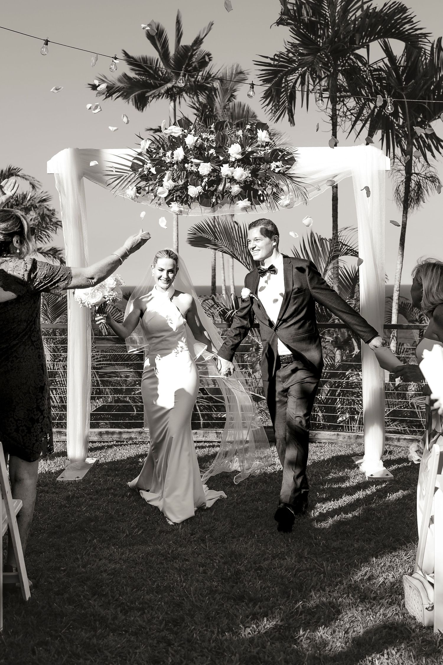 Black and white photo of a joyful newlywed couple exiting their outdoor wedding ceremony under a floral arch.