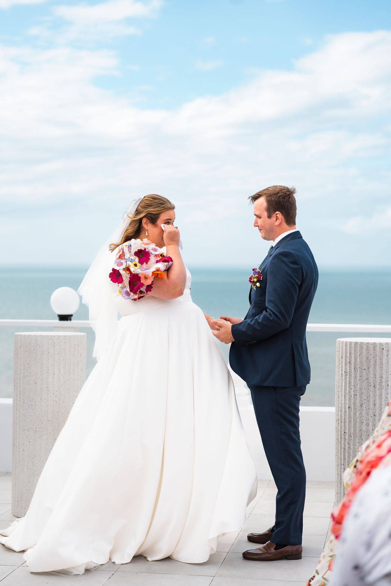 Bride and groom exchange vows on oceanfront rooftop terrace, bride holding vibrant pink and red bouquet.