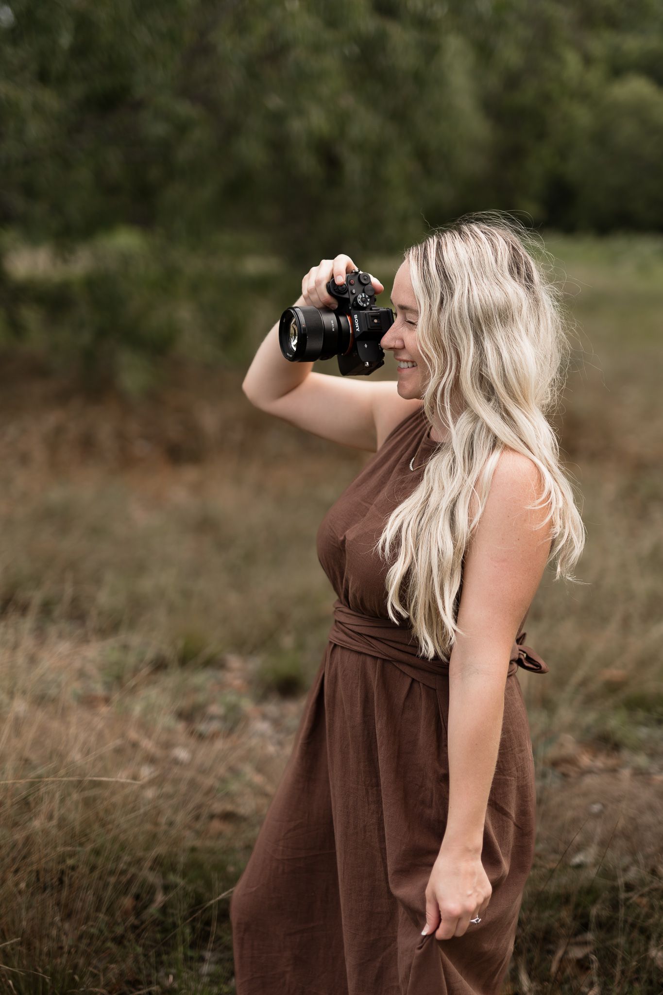 Blonde female photographer in brown dress smiling while taking photos in a grassy outdoor field.