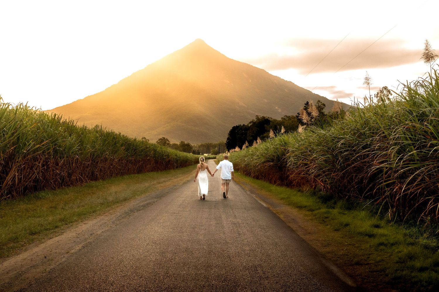 Couple walking hand in hand down a road toward a sunlit mountain surrounded by lush green fields at sunset.