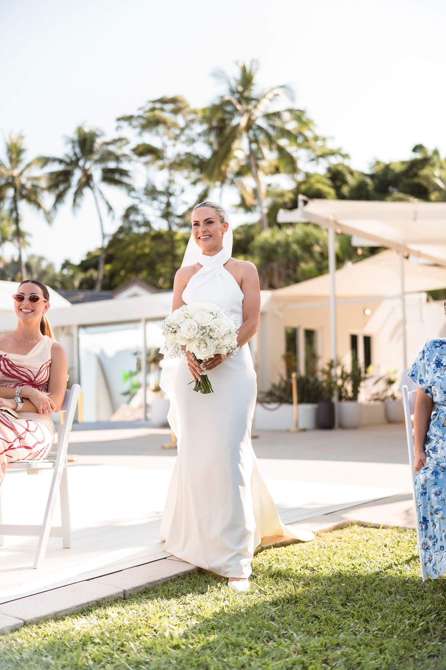 Smiling bride in white halter gown walks down outdoor aisle holding white bouquet with palm trees in background.