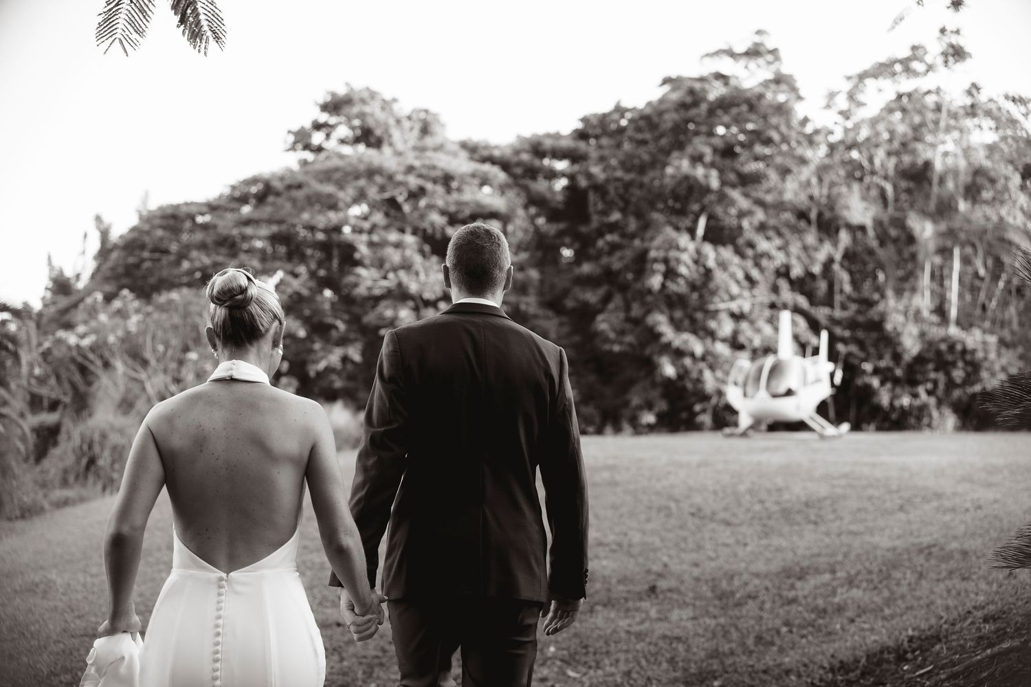 Black and white photo of bride and groom walking hand in hand toward a helicopter on their wedding day.