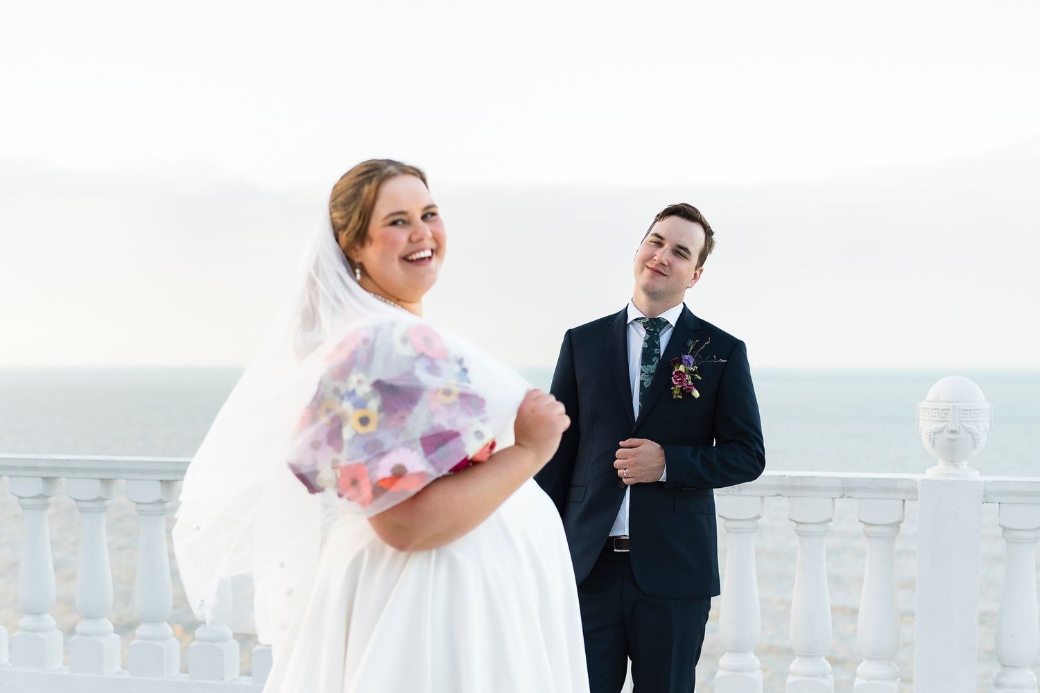 Bride and groom laughing together on a balcony overlooking the ocean on their wedding day.