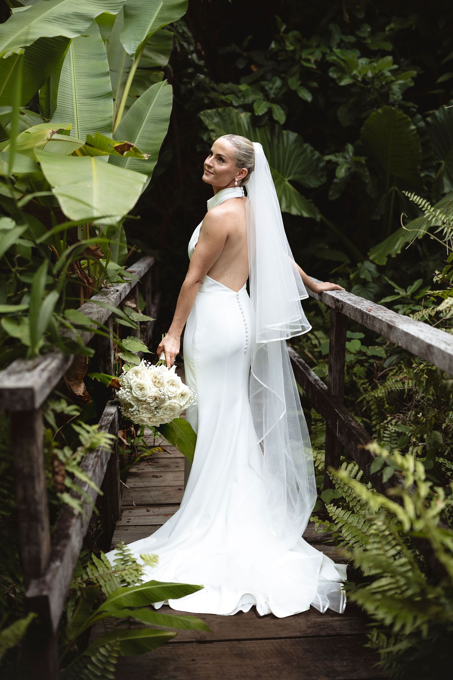 Elegant bride in backless white gown with veil on a wooden bridge.