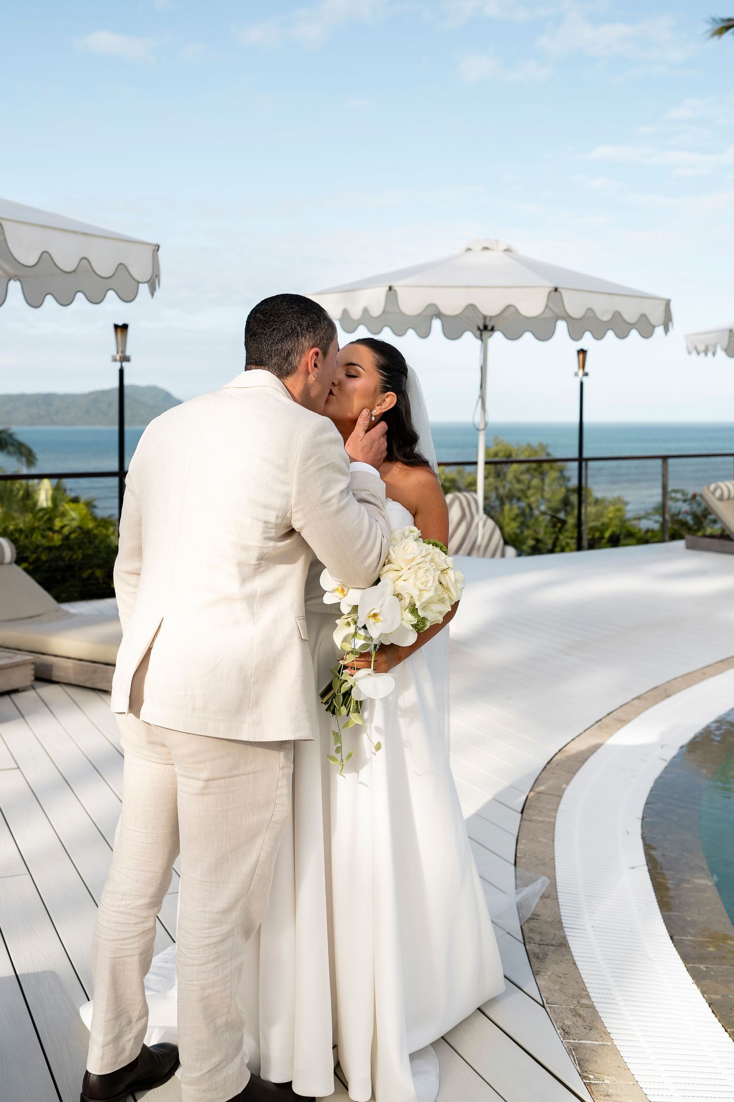 Bride and groom sharing a kiss at outdoor wedding venue with ocean view and white umbrellas.