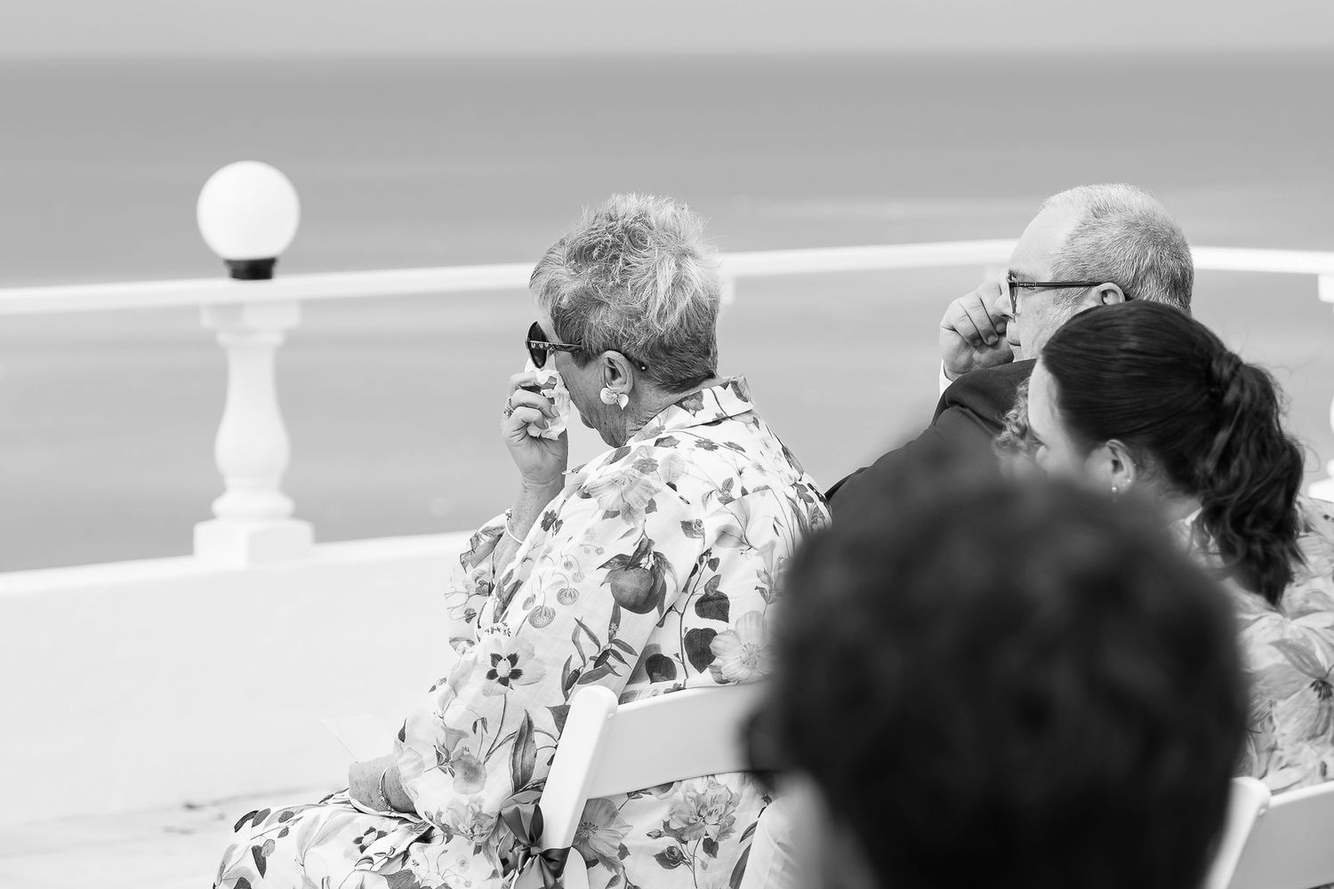 Black and white photo of elderly people sitting outdoors near the ocean, one talking on phone.