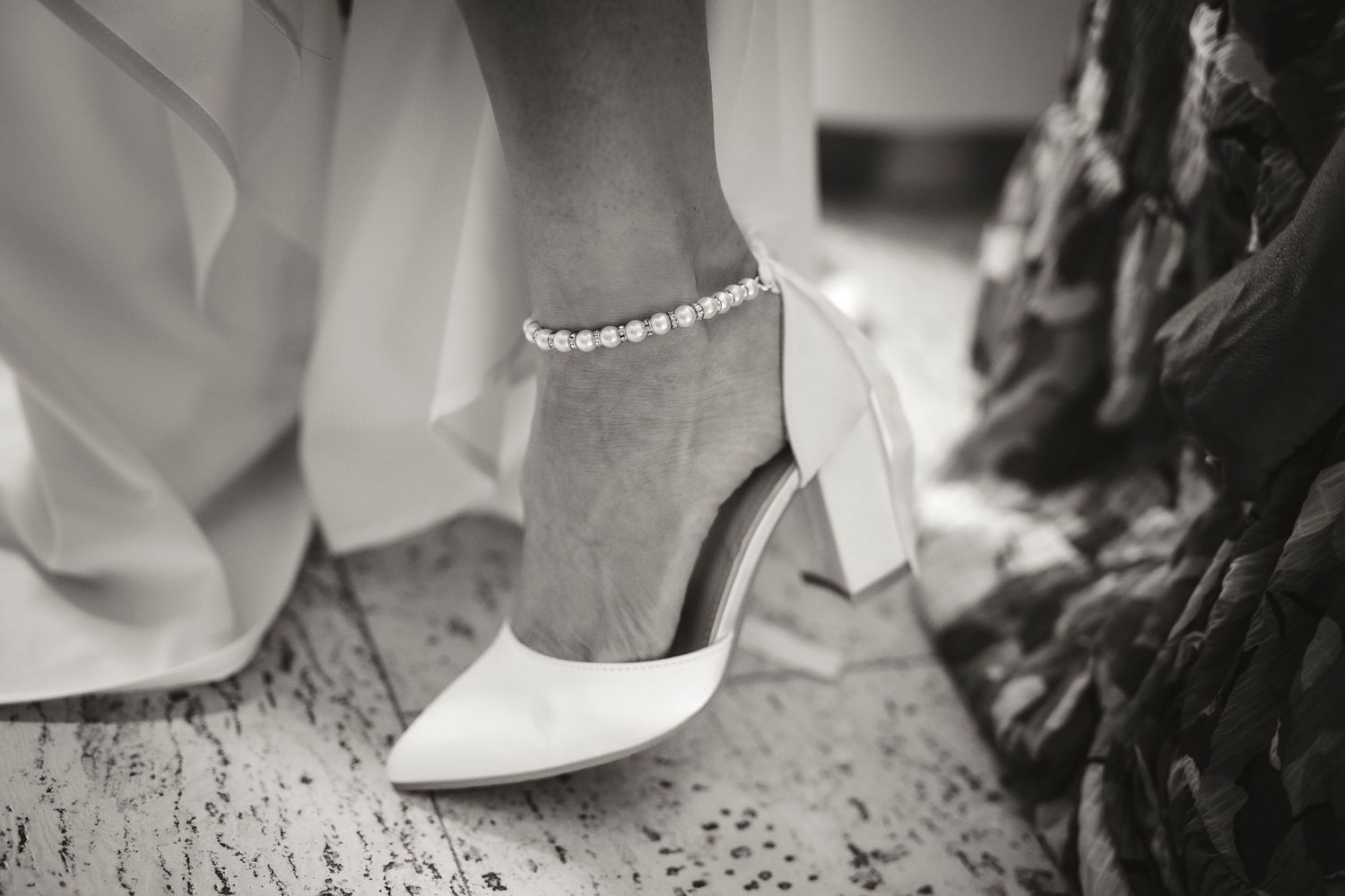 Close-up of a bride wearing white satin block heel shoes with a delicate pearl anklet on her wedding day.