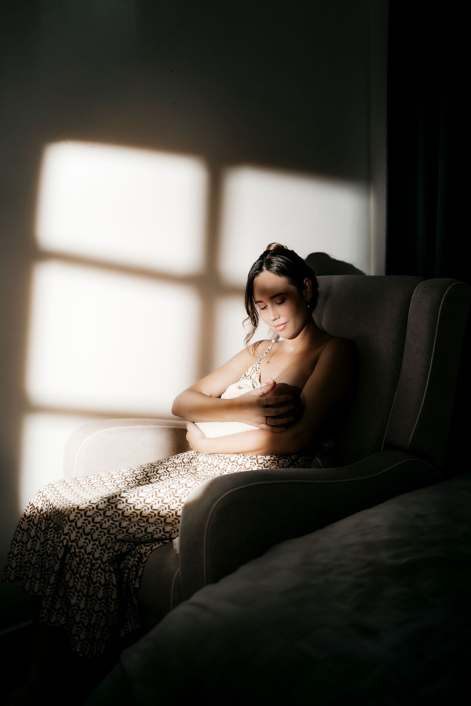 Woman sitting in armchair holding newborn baby in soft window light.