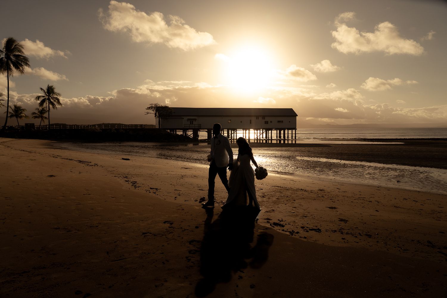 Silhouette of bride and groom walking on beach at sunset with pier in background.