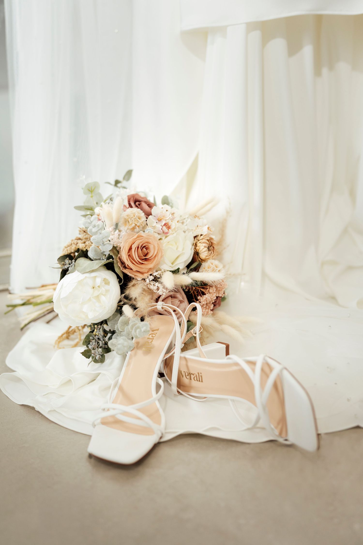 Bridal bouquet of roses and white flowers beside elegant white strappy heels on a soft white fabric backdrop.