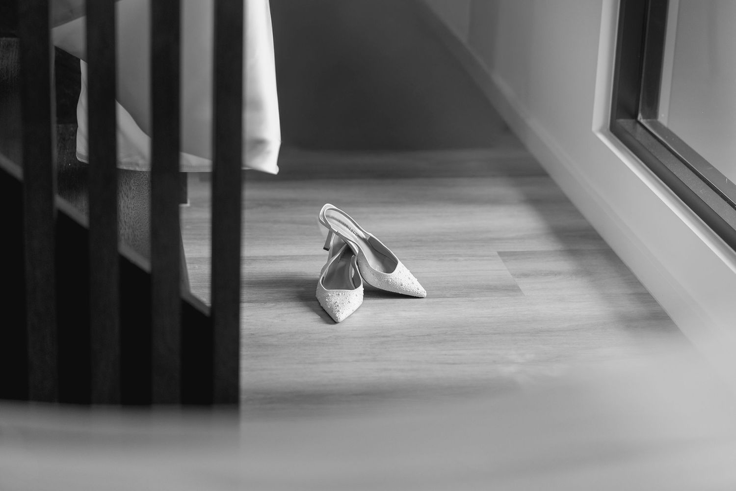 Black and white photo of elegant white high heel shoes placed on a wooden floor near a window and staircase.