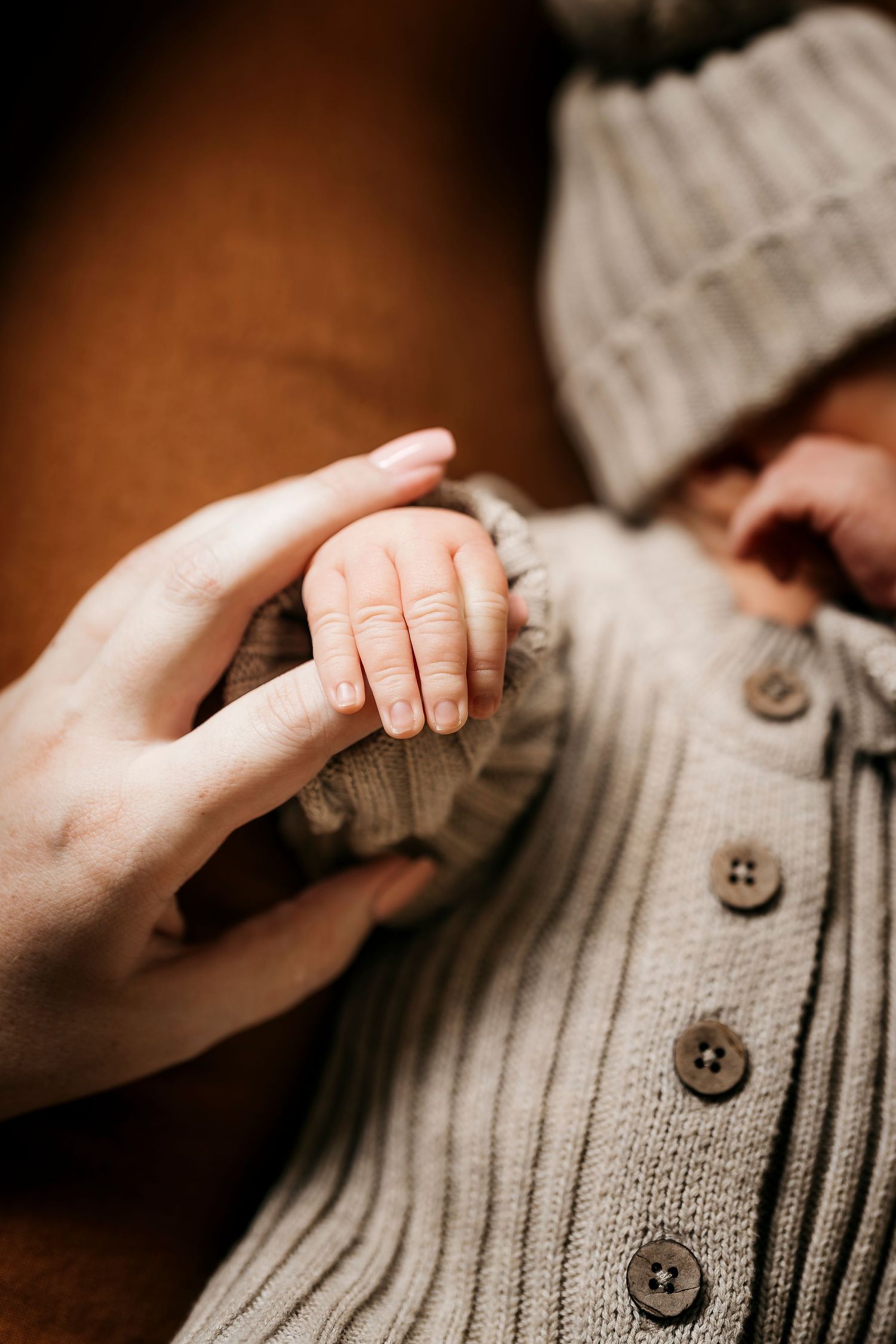 Close-up of newborn baby gripping parent's finger wearing knit grey outfit and hat.