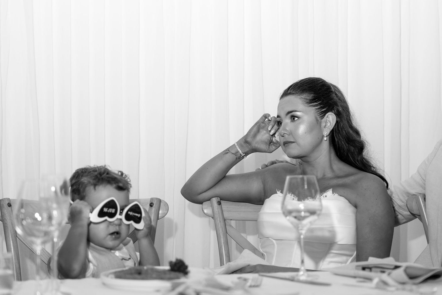 Black and white photo of a woman wiping tears at a wedding reception while a toddler wears novelty sunglasses at the table.