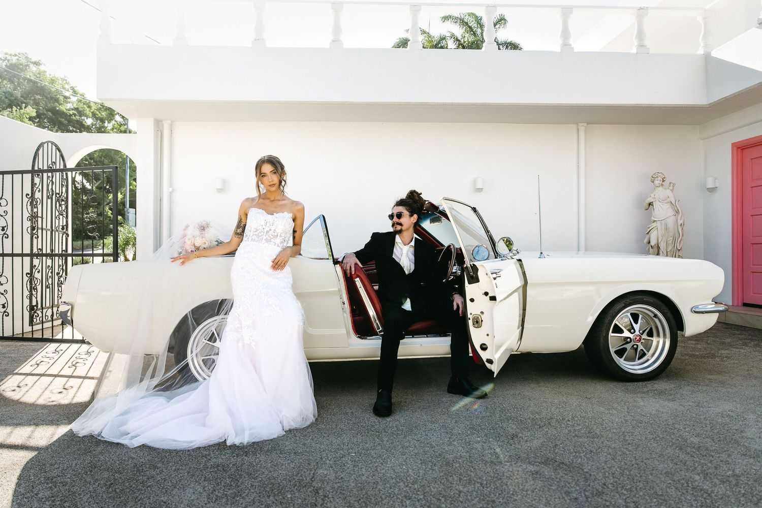 Bride and groom posing beside a classic white vintage car outside a modern white building on their wedding day.