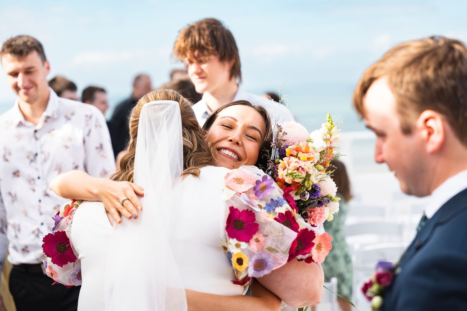 Bride hugging a guest while holding a colorful bouquet at an outdoor beach wedding ceremony.